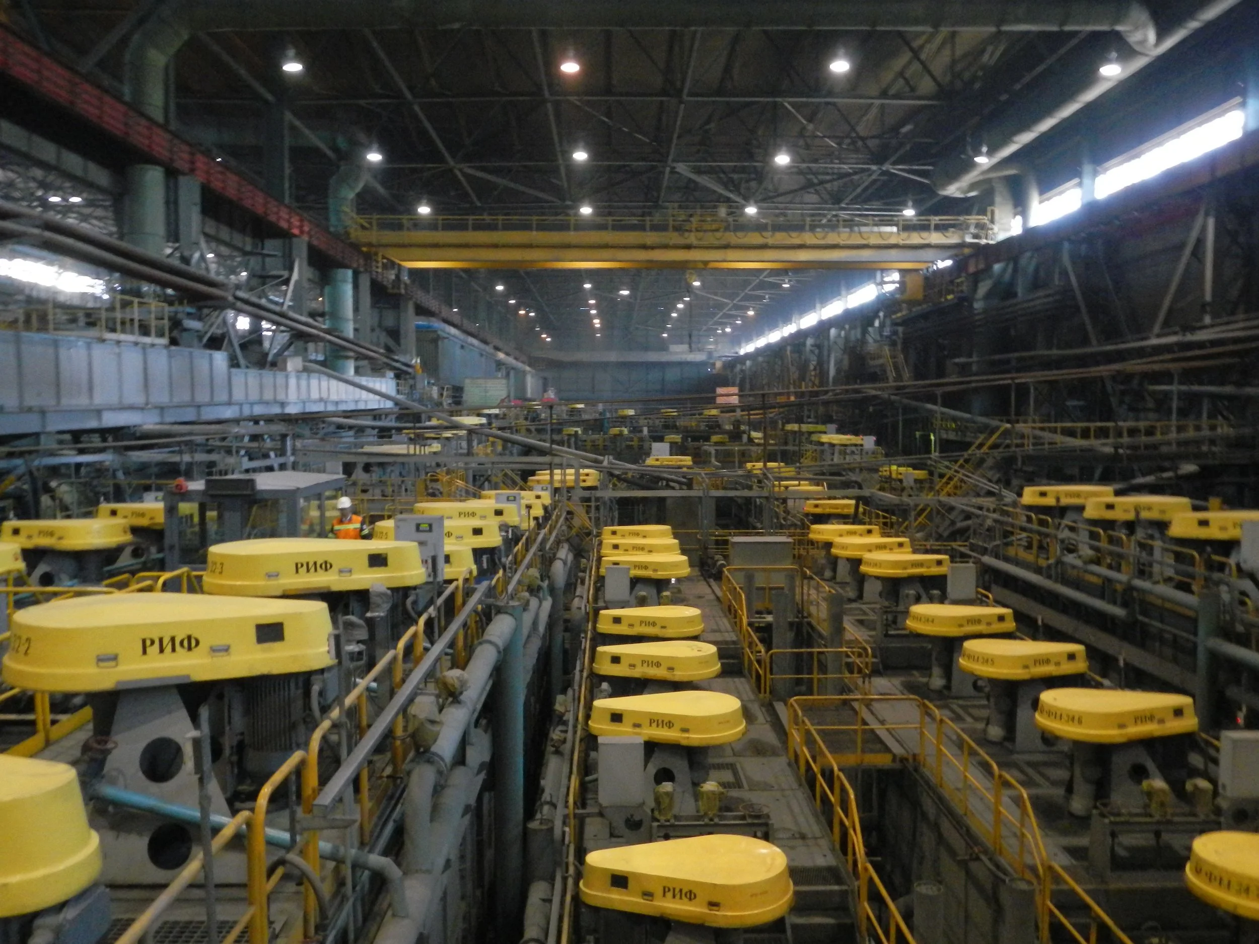 Interior of an industrial factory with yellow machinery and overhead crane, workers in safety vests and helmets, pipes, and metal framework.