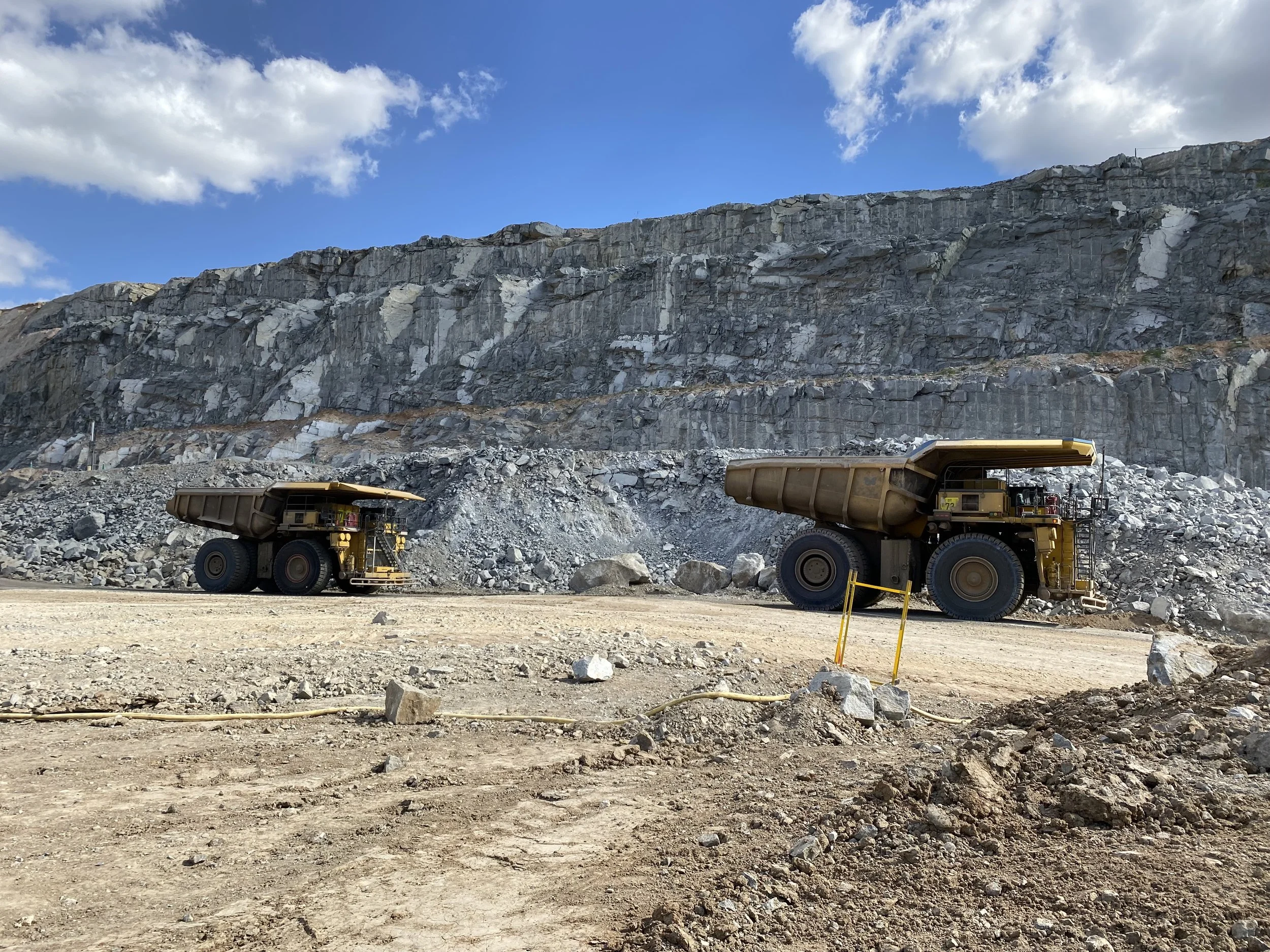 View of a large rocky quarry or mining site with two yellow haul trucks in the foreground, gray rock face in the background, and blue sky with white clouds overhead.
