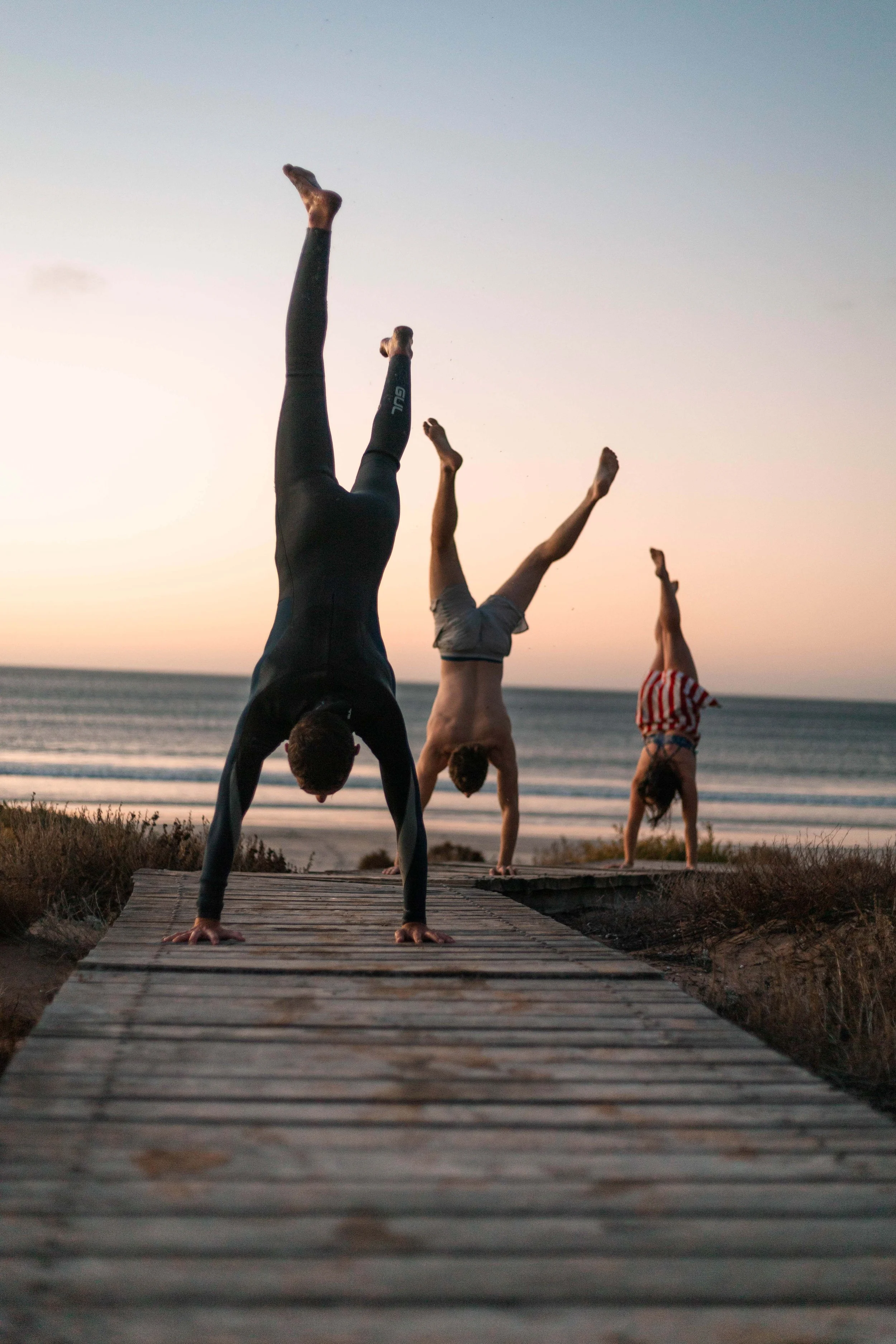 A group of people practicing yoga or handstands on a wooden pathway at the beach during sunset.