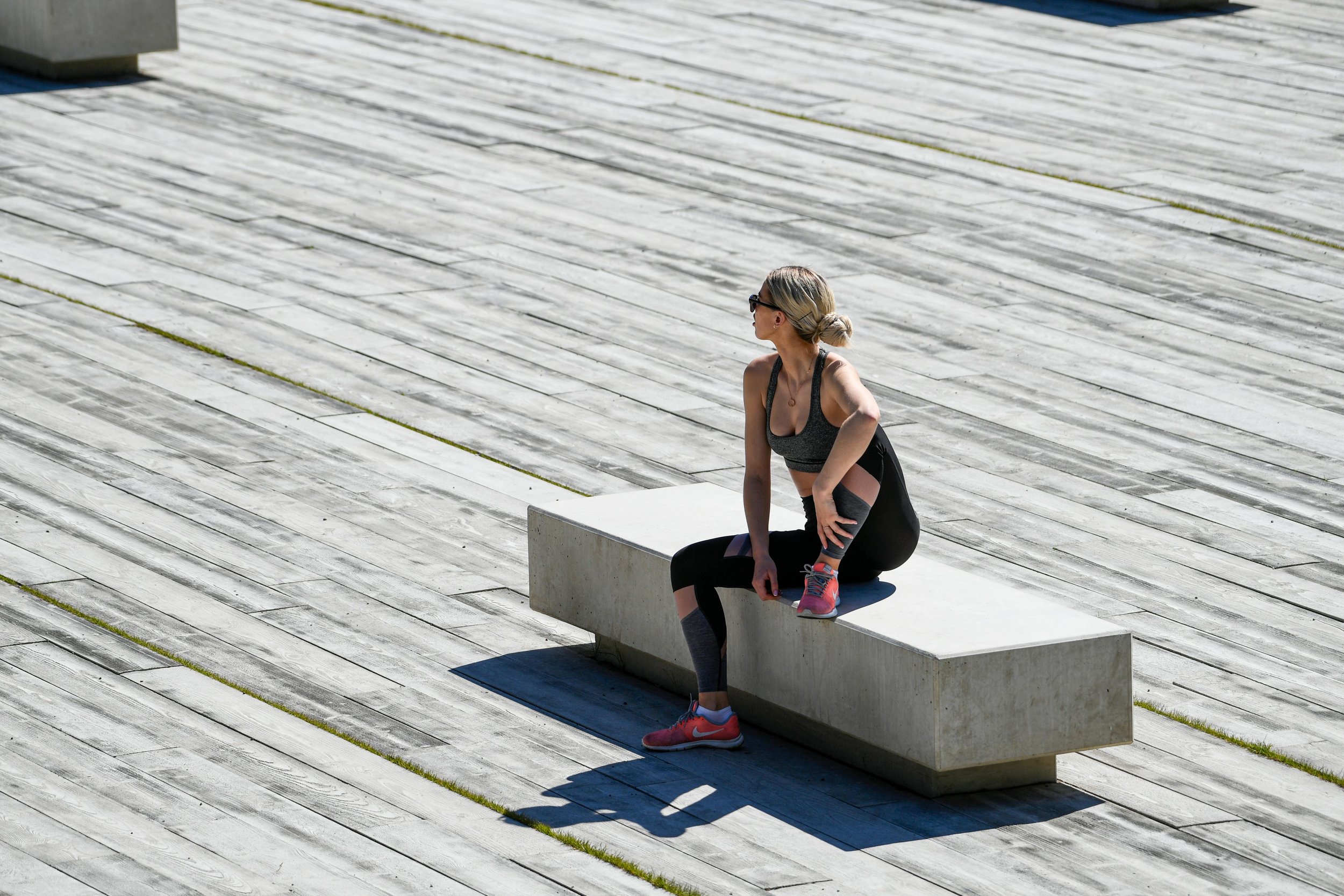 A woman in athletic clothing and pink sneakers sitting on a concrete bench on a large wooden deck outdoors.