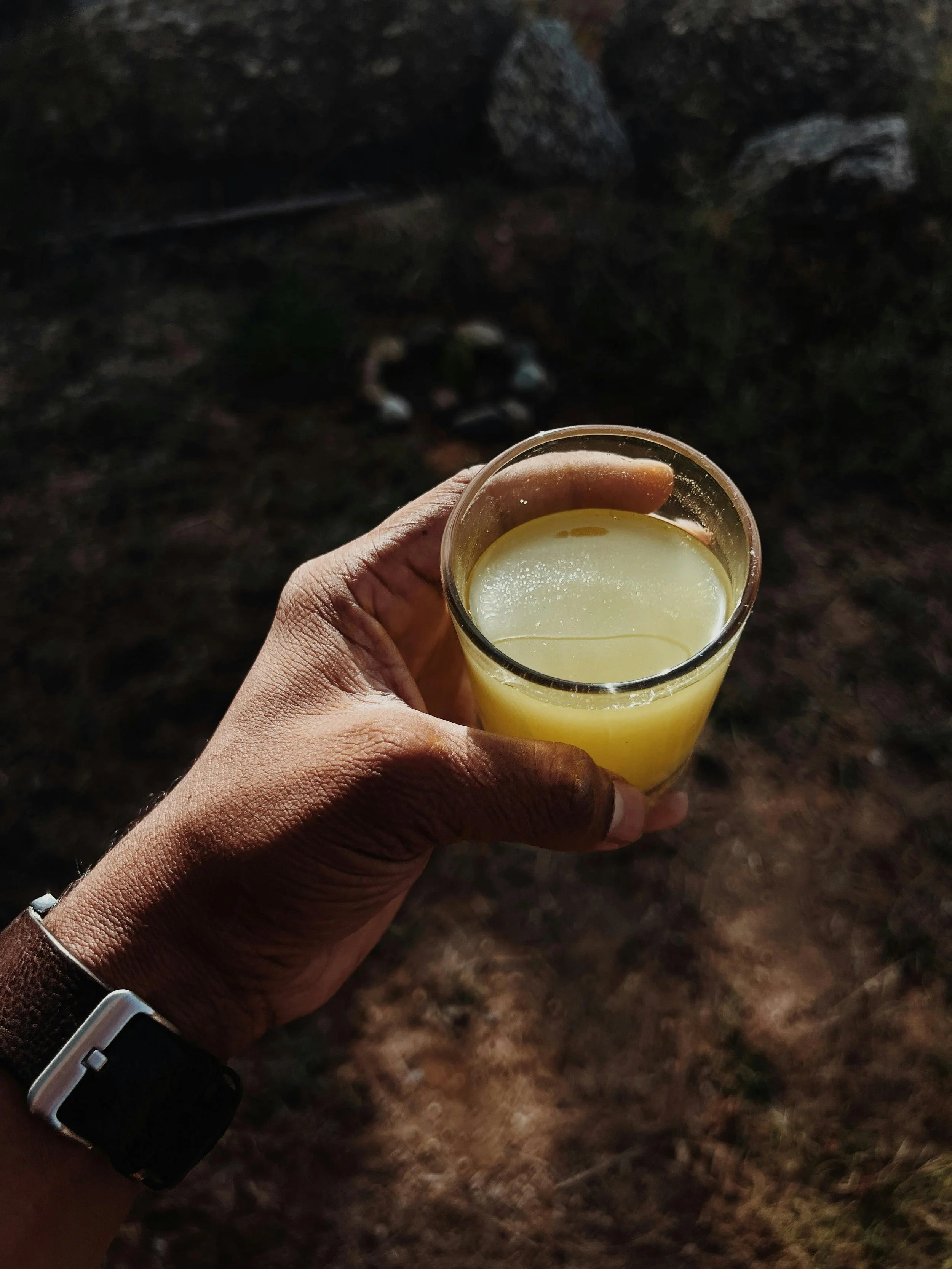Hand holding a glass of yellowish liquid, dark background with rocks and soil.