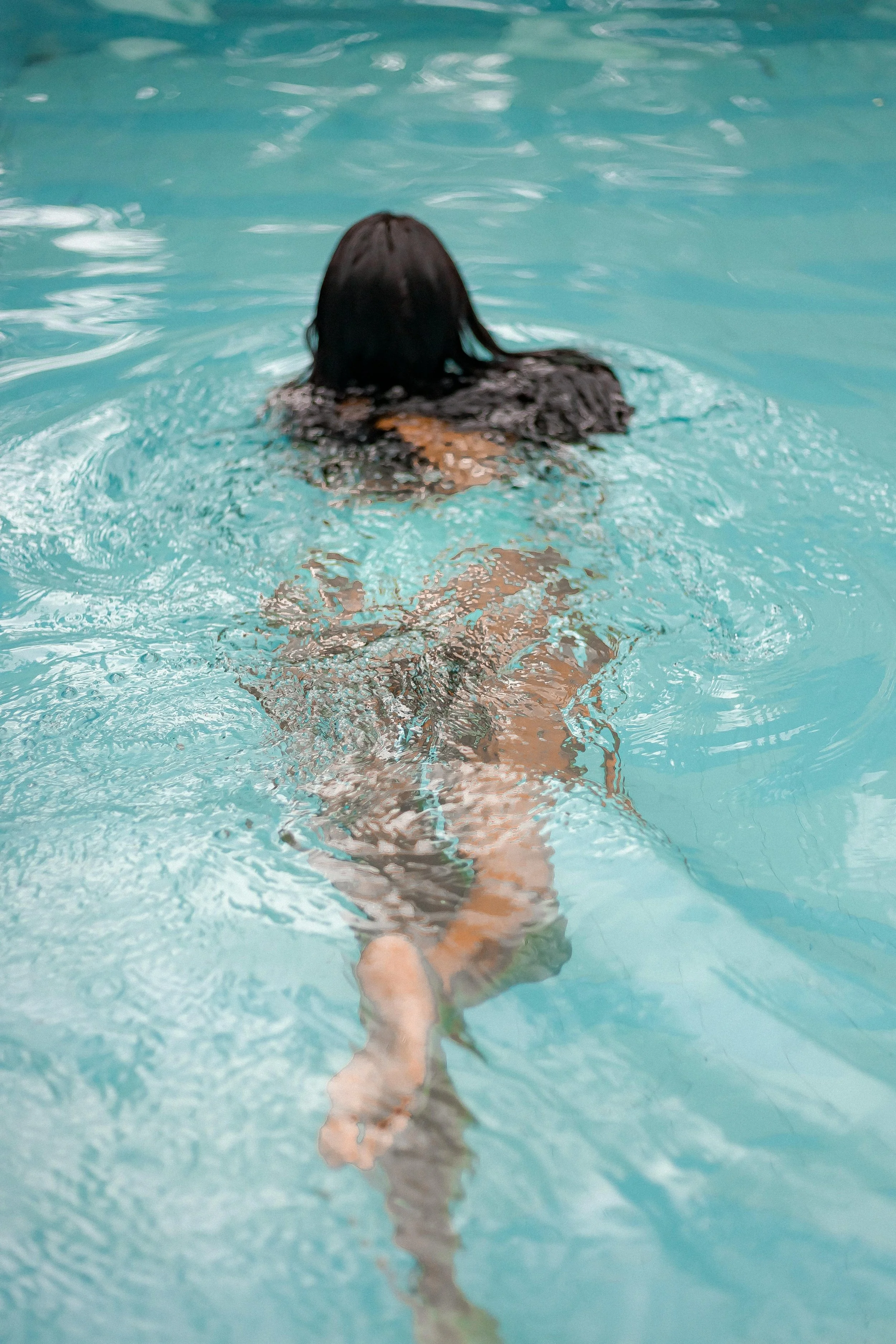 A person with dark hair swimming underwater in a pool, with their head and upper body submerged and their legs slightly bent.