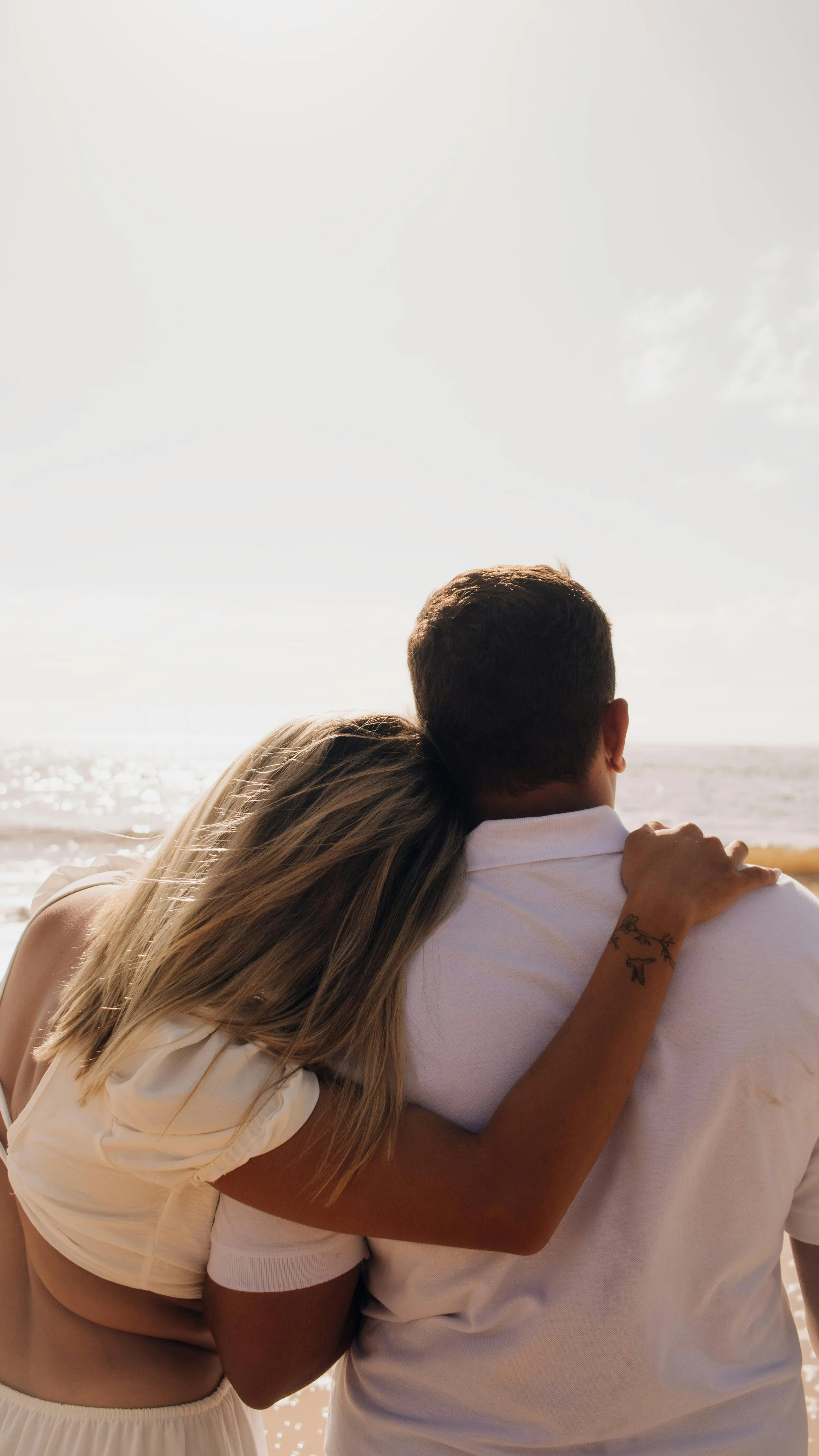 A couple embracing on a beach, with the woman resting her head on the man's shoulder, looking out at the ocean.