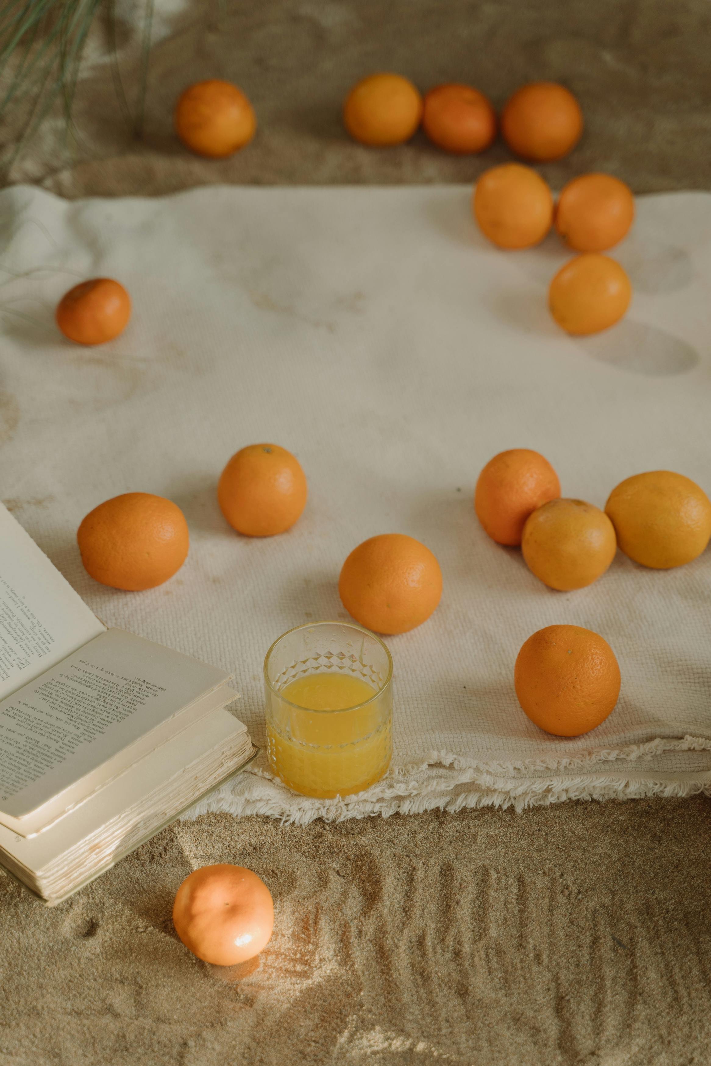 A table setting with several oranges, an open book, a glass of orange juice, and a white cloth on a textured surface.