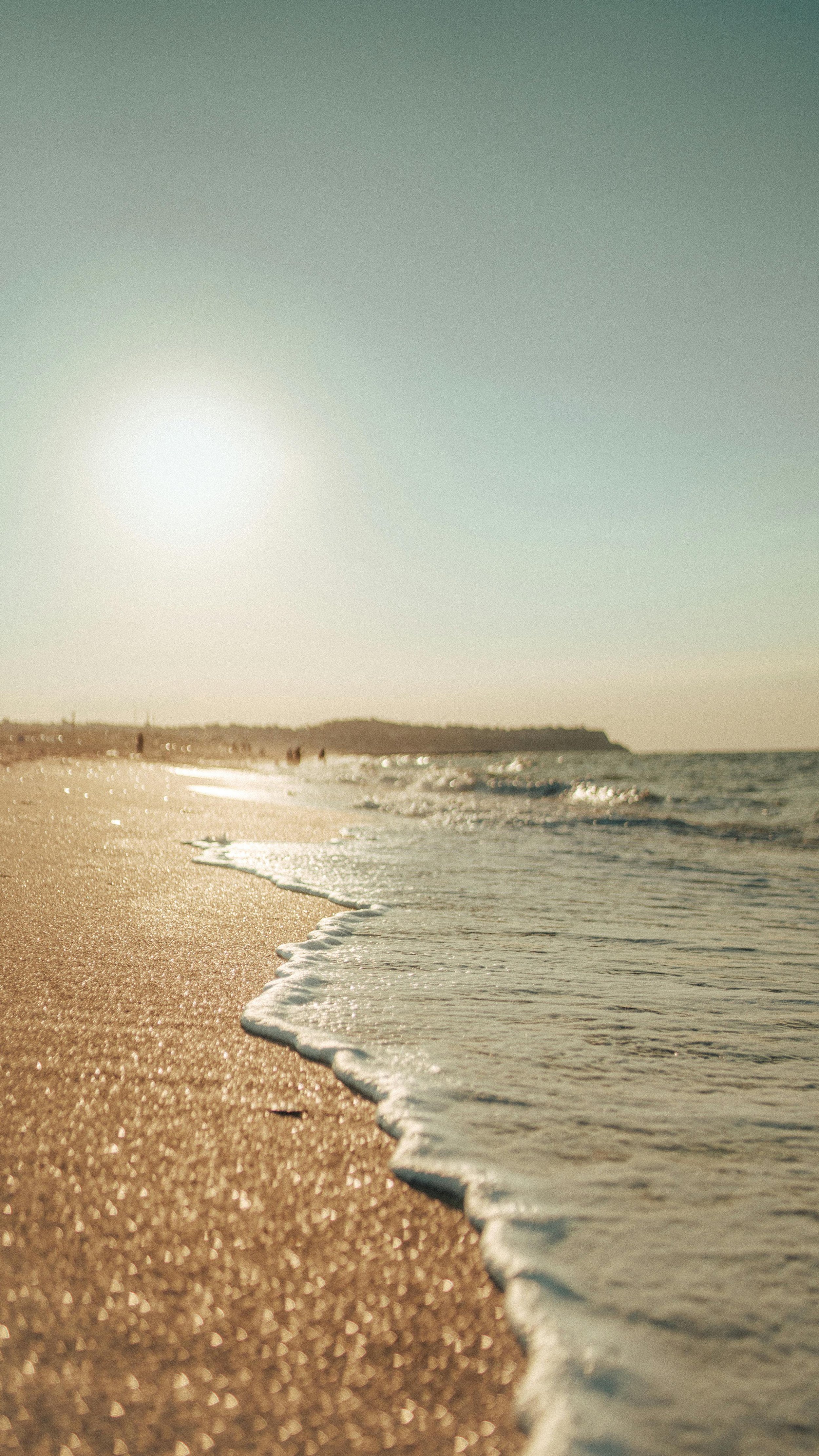 Sunset over a sandy beach with gentle waves in the foreground and distant figures walking along the shoreline.