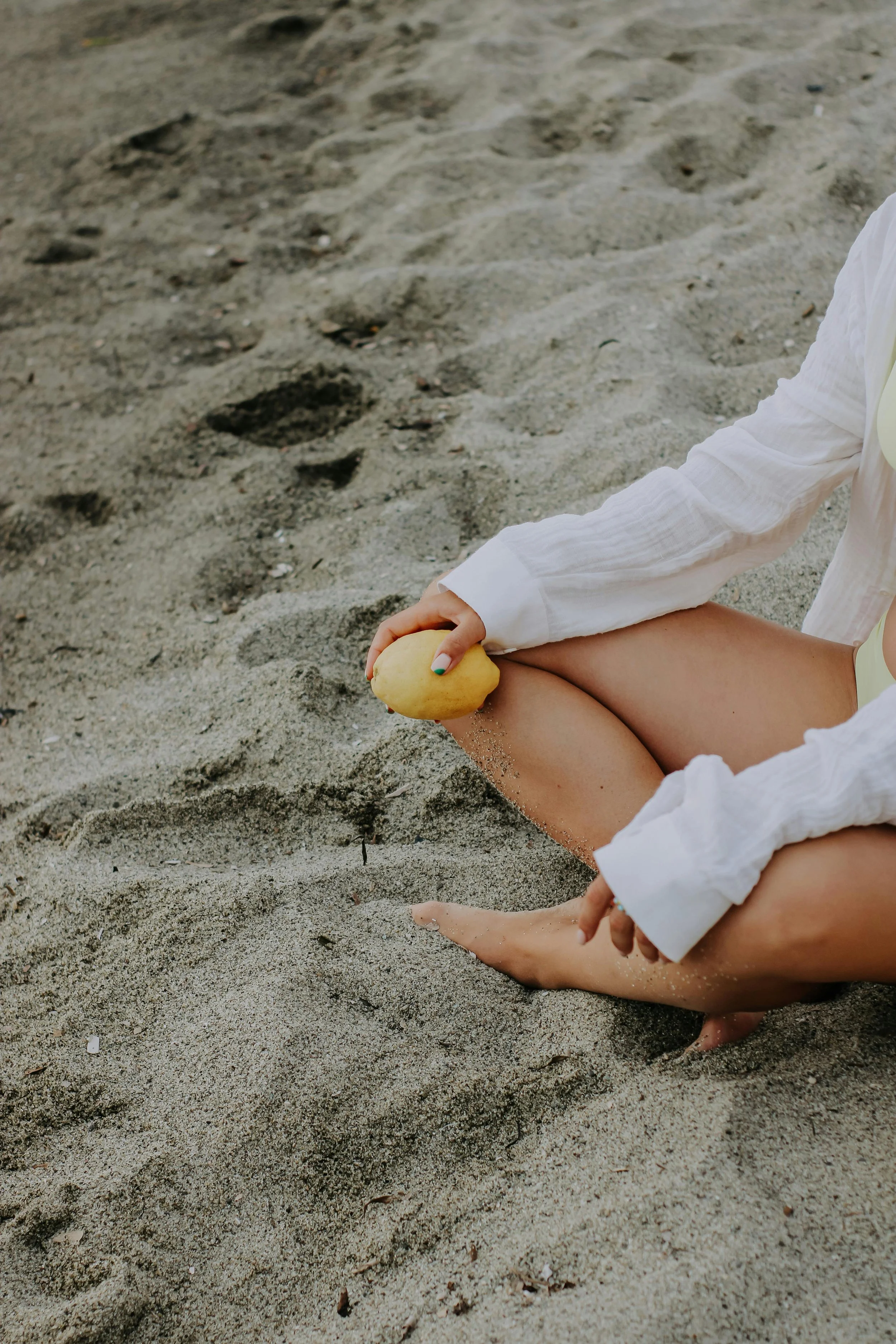 Person sitting on the sand at the beach, holding a yellow object in their hands, wearing a white long-sleeve shirt with a pale yellow inner shirt visible. The person's legs are crossed and sandy beach is seen around them.