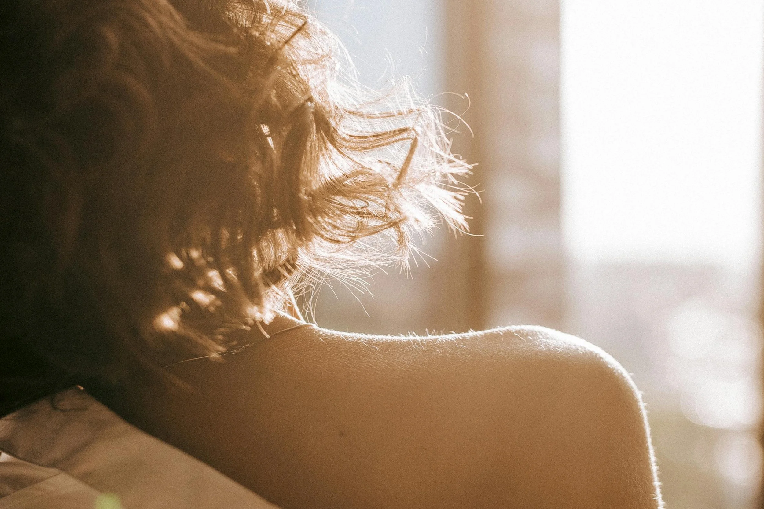 A close-up of a person's shoulder and curly hair, illuminated by soft sunlight coming from the right side of the image.