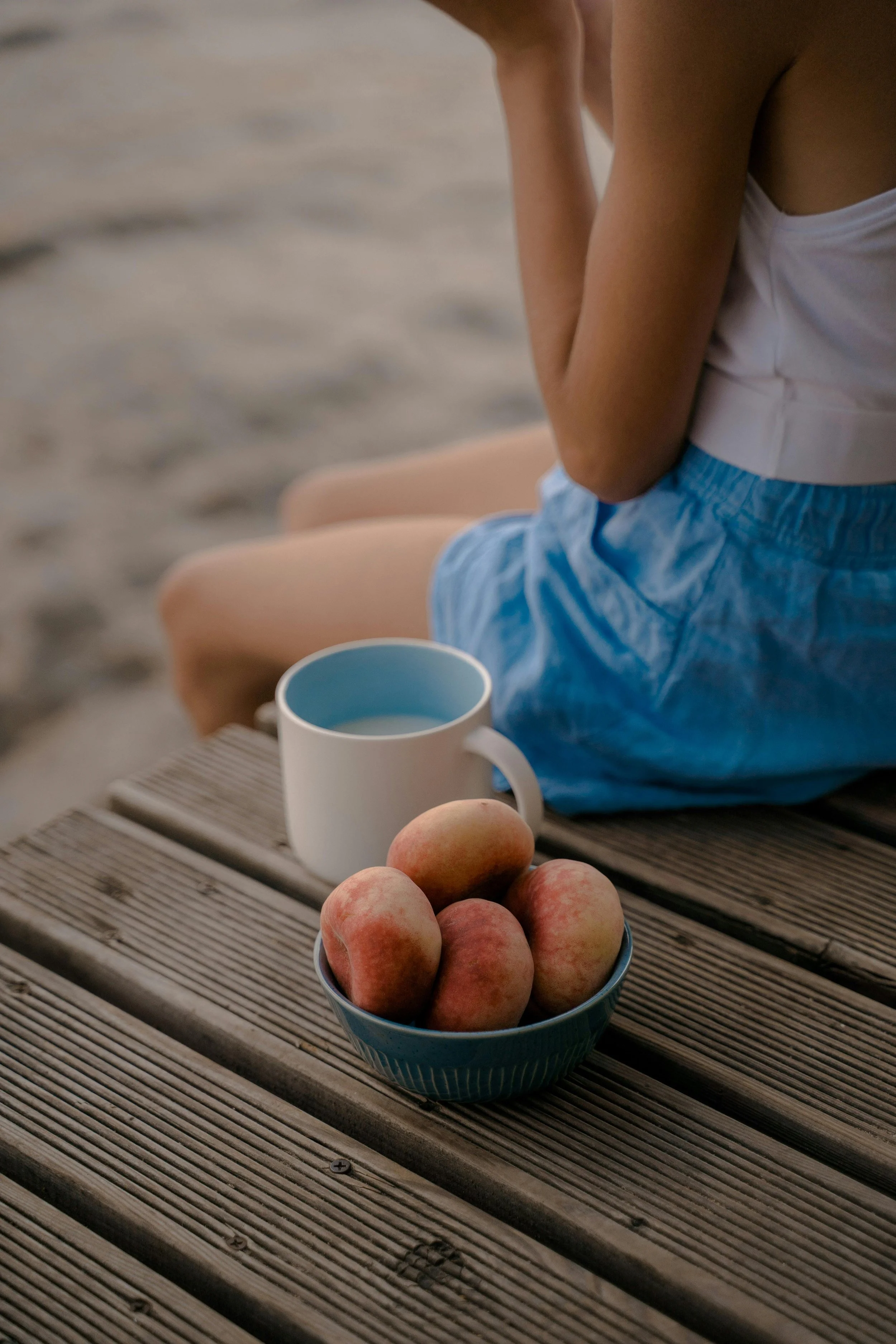 A bowl of peaches on a wooden table with a person sitting nearby wearing a white tank top and blue shorts, a white mug, and a striped table mat.