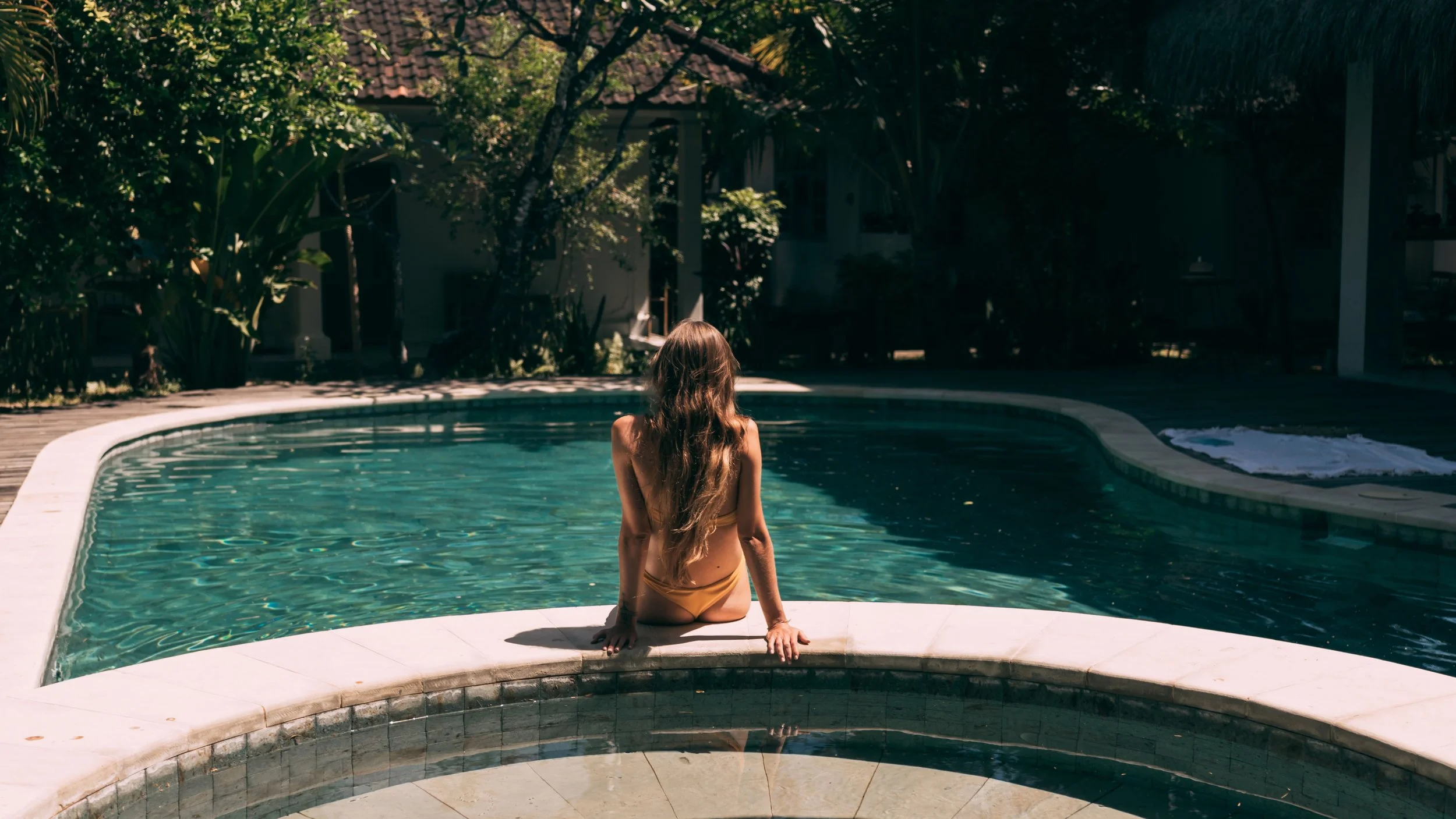 A woman with long hair sitting on the edge of a swimming pool, facing away from the camera, surrounded by lush greenery and a house in the background.