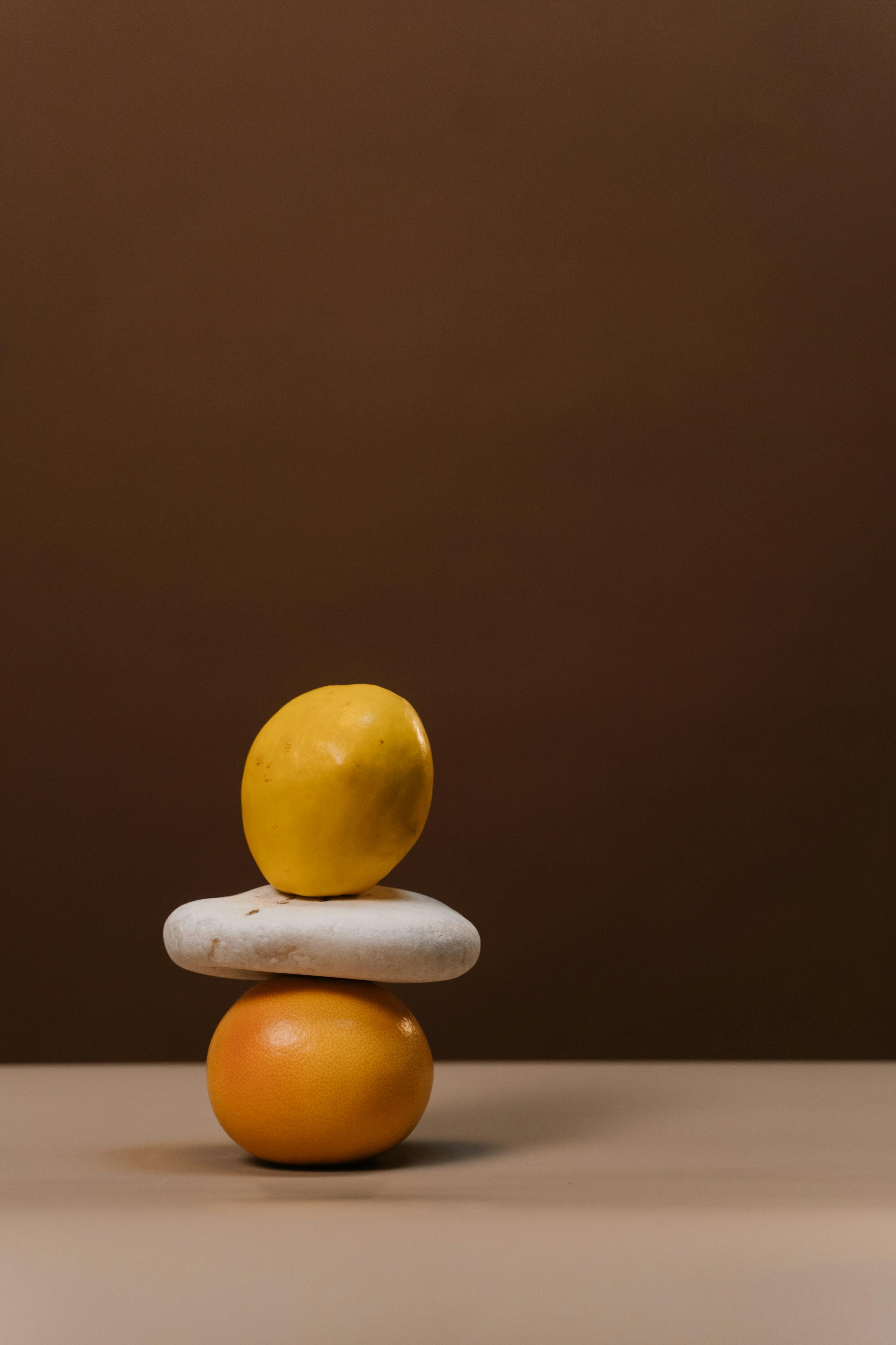 Stacked fruits with a yellow apple on top, followed by a white stone, an orange, and a yellow grapefruit, against a dark brown background.