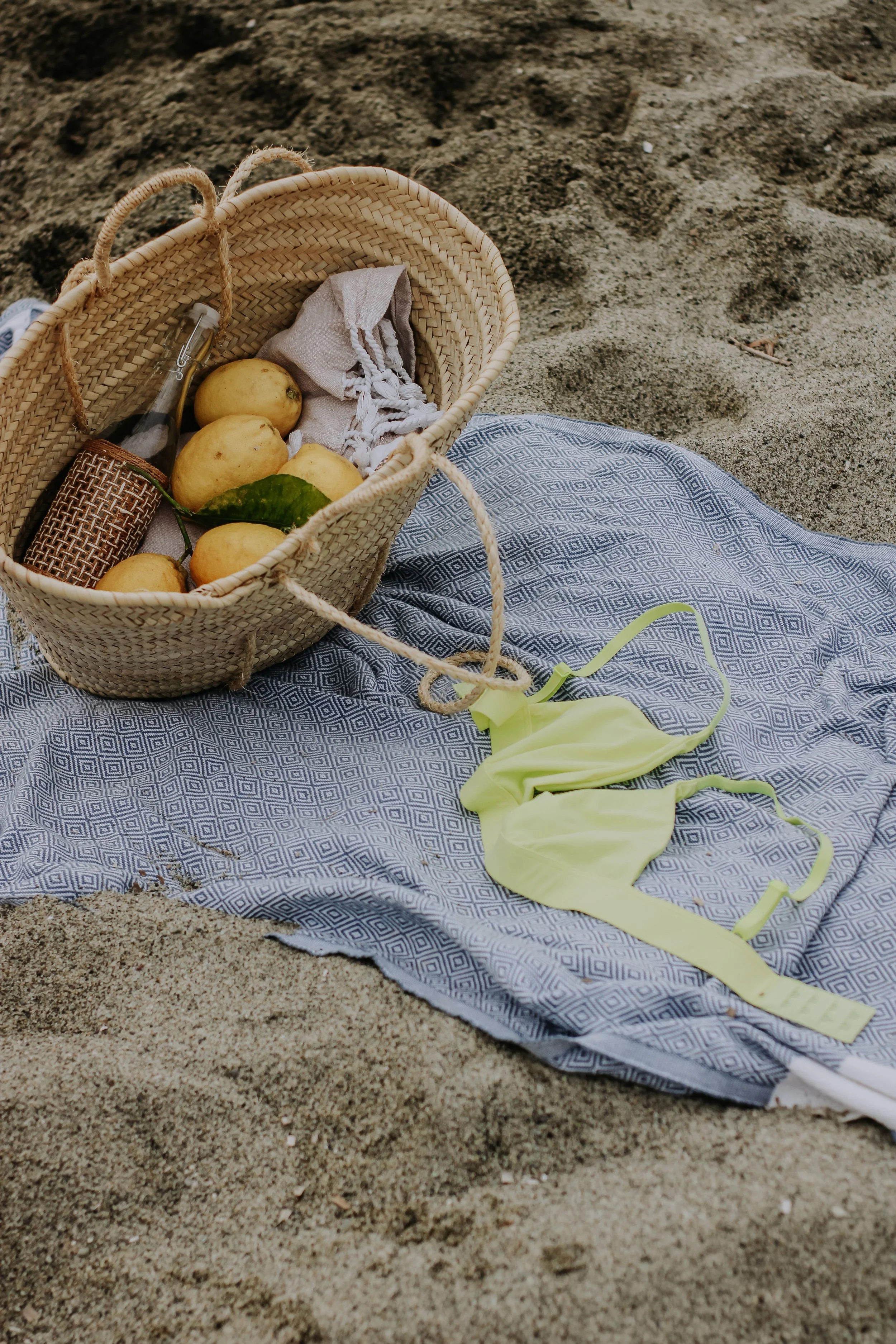 A picnic basket on sandy ground with a blue patterned cloth, yellow balloons, a yellow ribbon, and a small glass bottle inside.