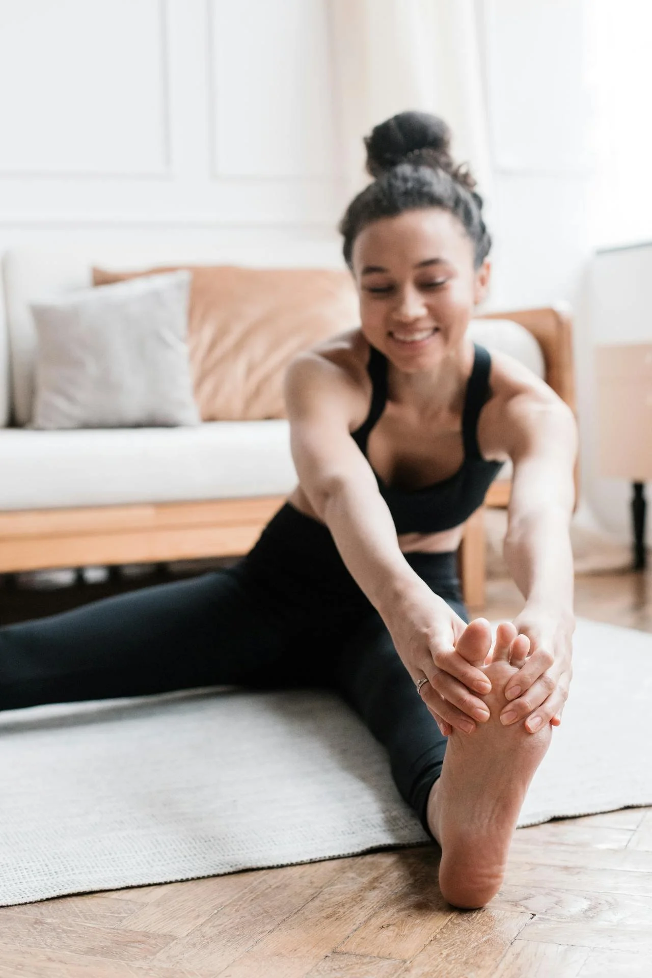 A woman with dark hair in a bun, smiling, practicing a seated forward bend stretch on a yoga mat in a well-lit room with a sofa and pillows in the background.