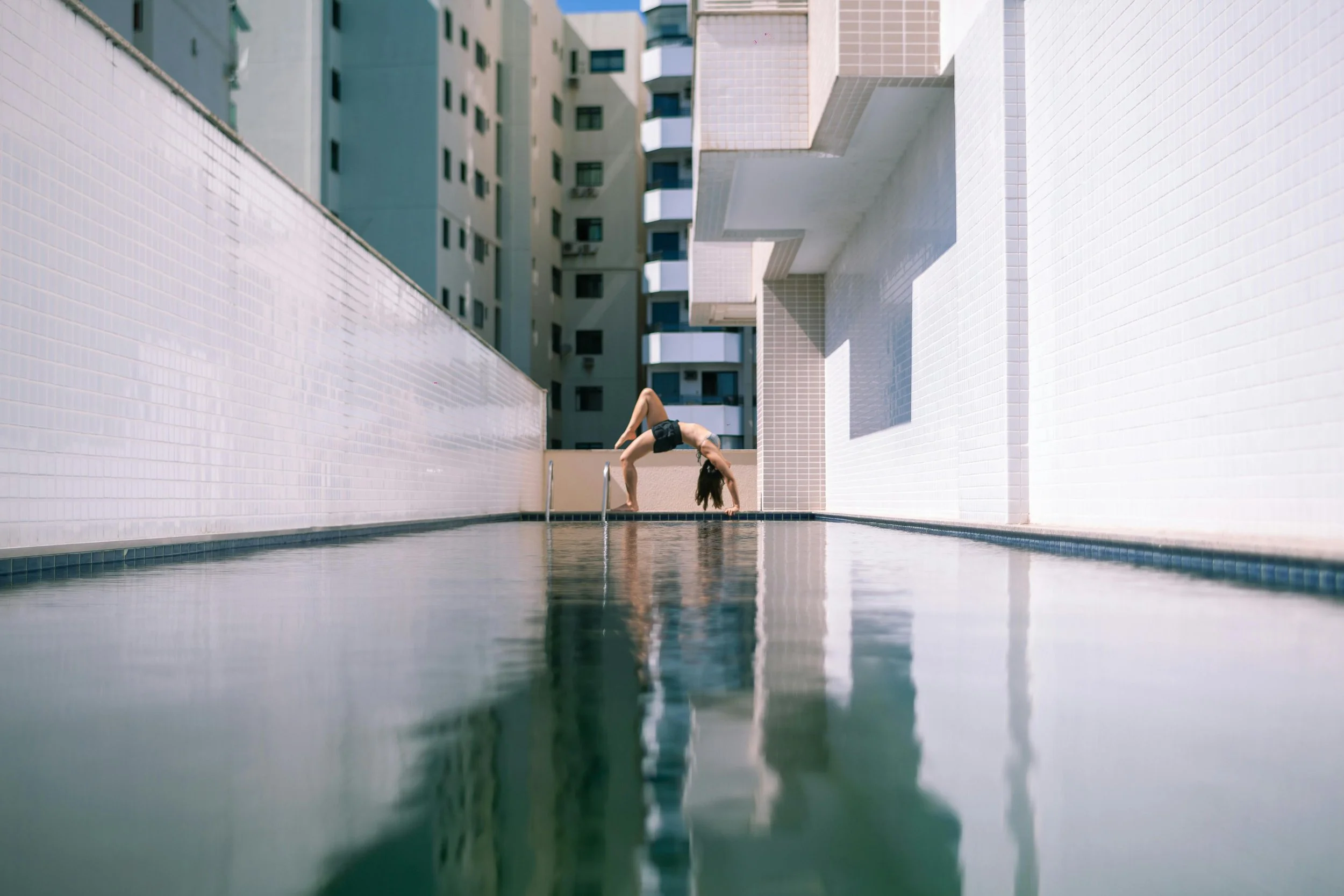 A woman in black athletic wear performing a backbend yoga pose on the edge of a rooftop pool with a city apartment building in the background.