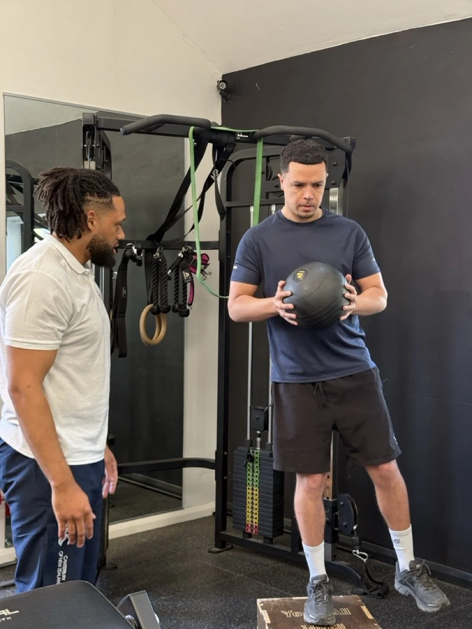 A man in a blue t-shirt and black shorts is holding a weight ball with a male physiotherapist in a white t-shirt observing him in a gym.