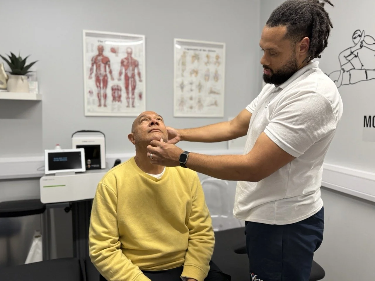 A physiotherapist examines a patient's neck in a consultation room with anatomical posters on the wall.
