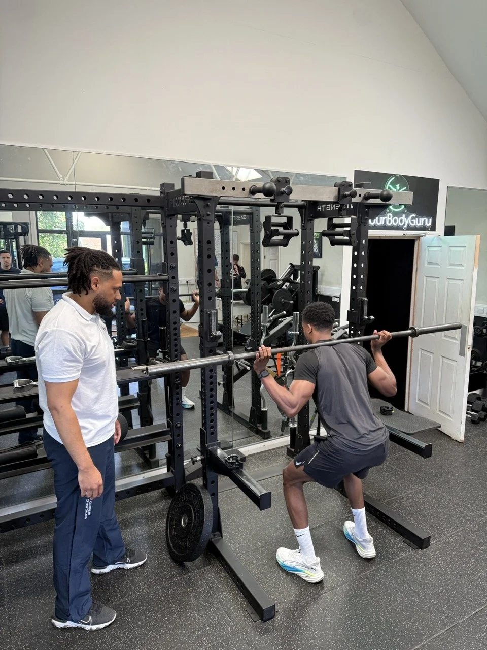 Man lifting a barbell on squat rack while a trainer watches in a gym.
