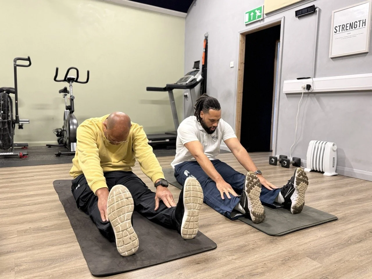 Two men are doing stretching exercises on exercise mats in a gym, sitting with legs extended and reaching for their toes.
