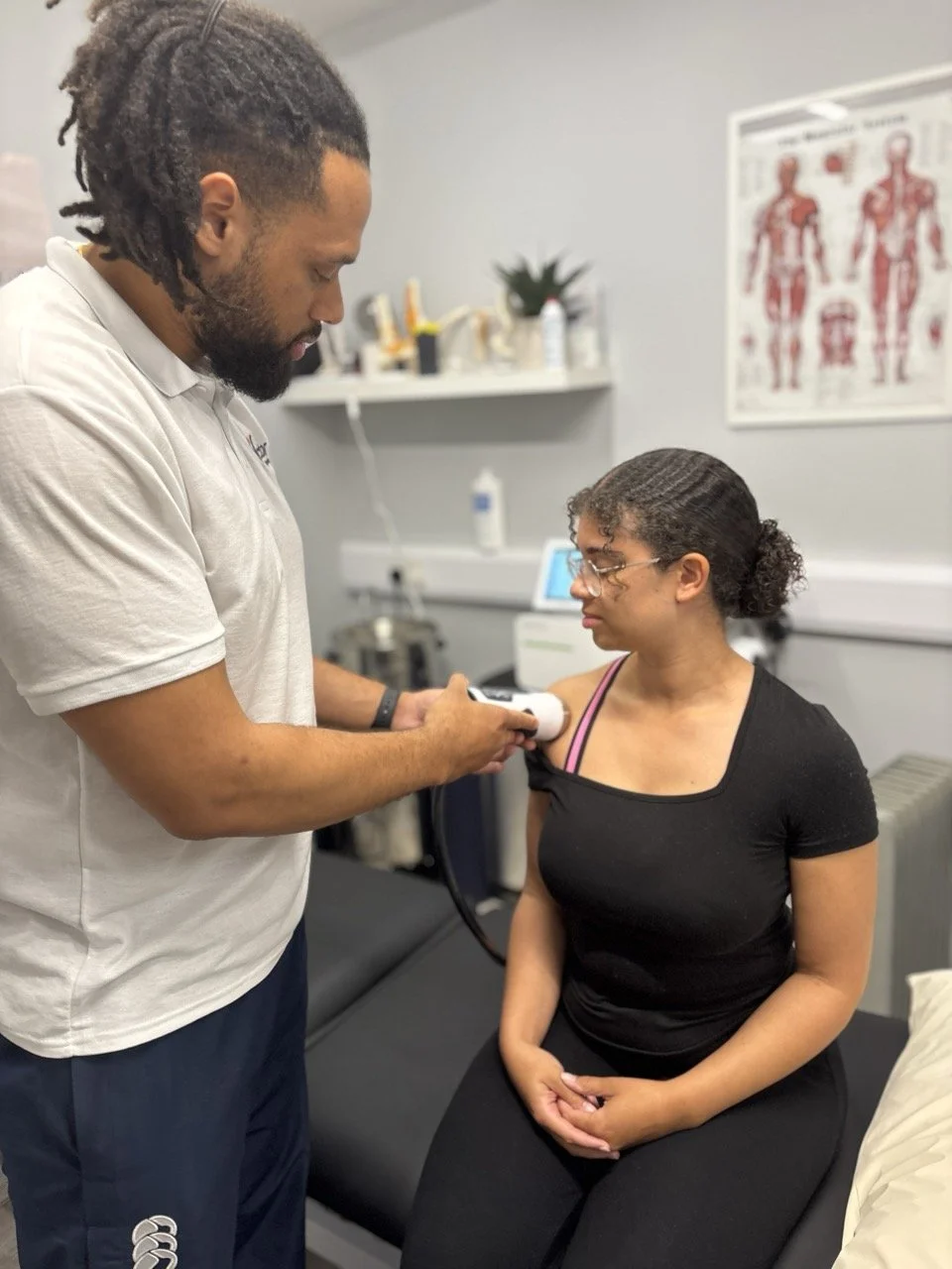 A healthcare professional uses a handheld ultrasound device on a woman's shoulder in a medical examination room.