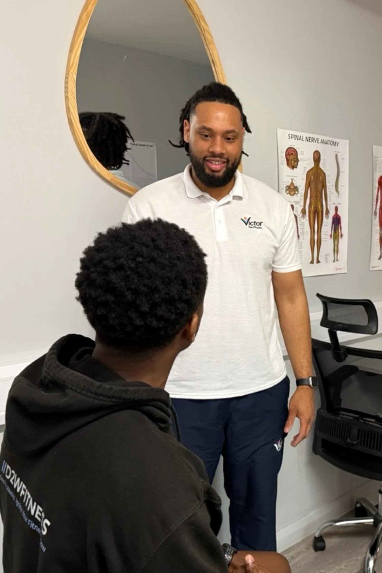 A man with a beard and dreadlocks wearing a white polo shirt is talking to a person in a black hoodie in a physio clinic room, with spinal nerve anatomy posters on the wall.