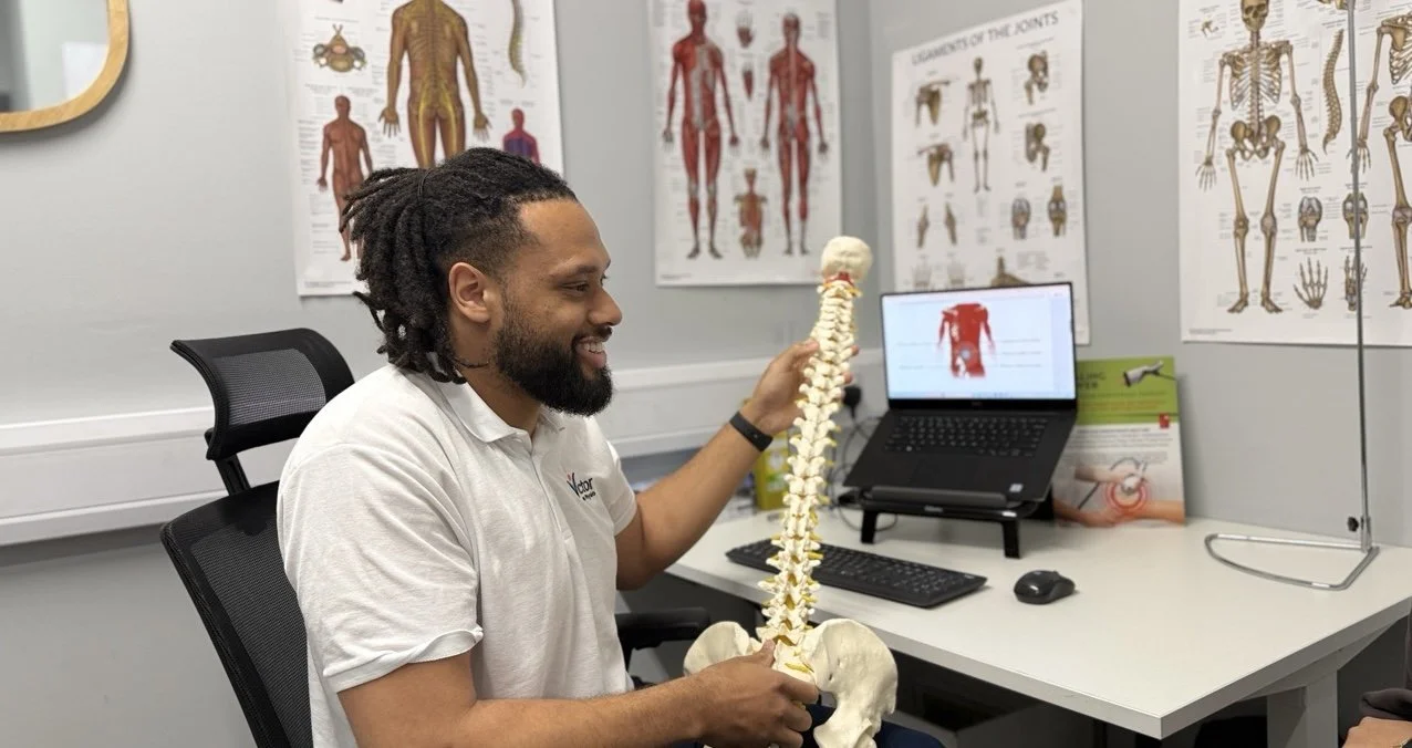 A man with dreadlocks smiling and holding a model of a human spine while sitting in a physiotherapy clinic room with anatomy posters on the wall and a laptop on the desk.