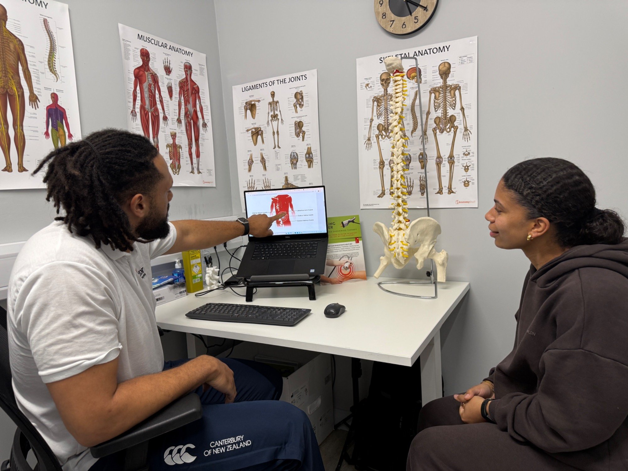 Two people in a physiotherapy clinic room with anatomy posters on the wall, a model of the spine on the table, and a physio pointing at a chart on a laptop screen.