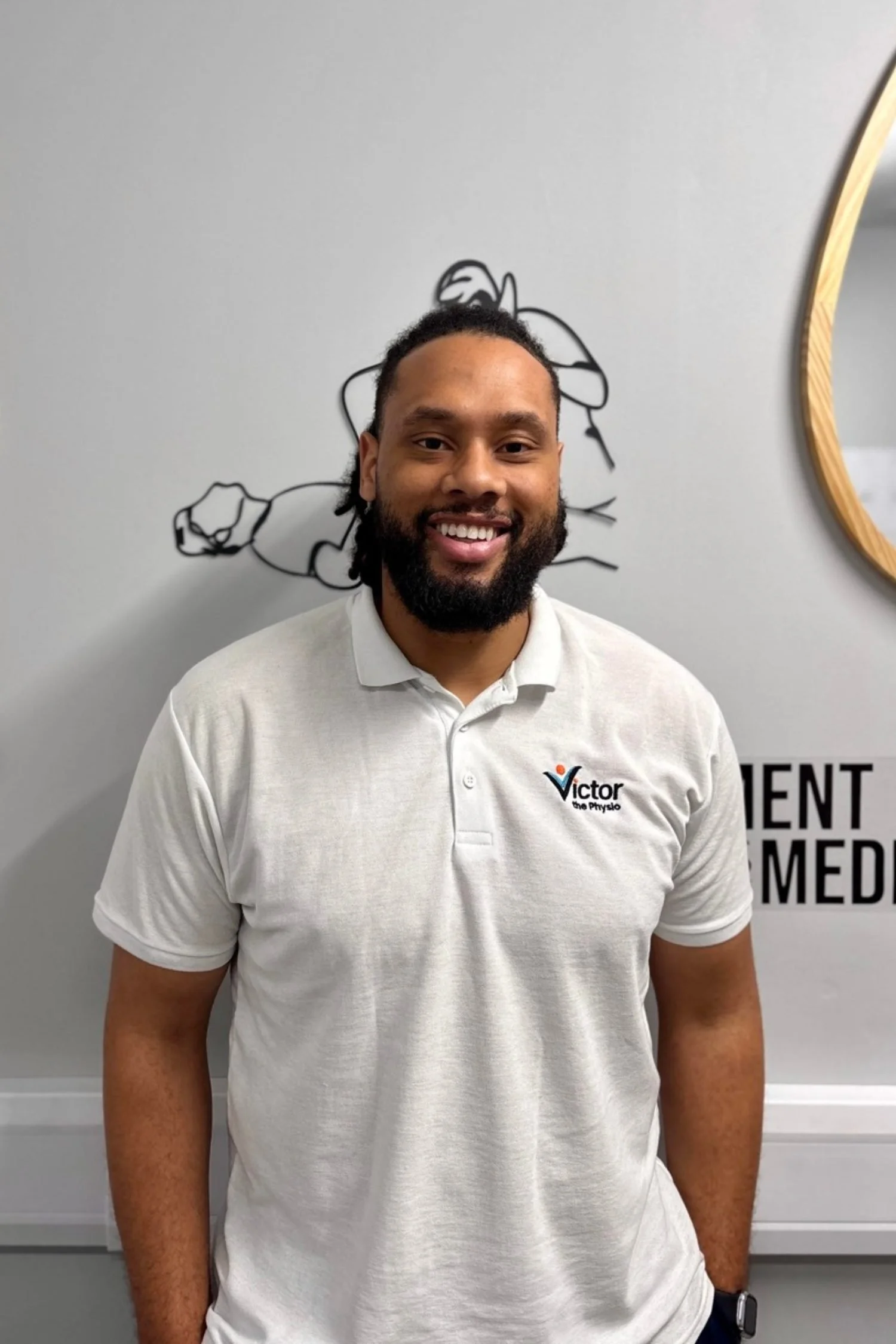 A smiling man with a beard and dreadlocks, wearing a white polo shirt with a logo, standing in front of a wall with a line art drawing of a person flexing muscles.