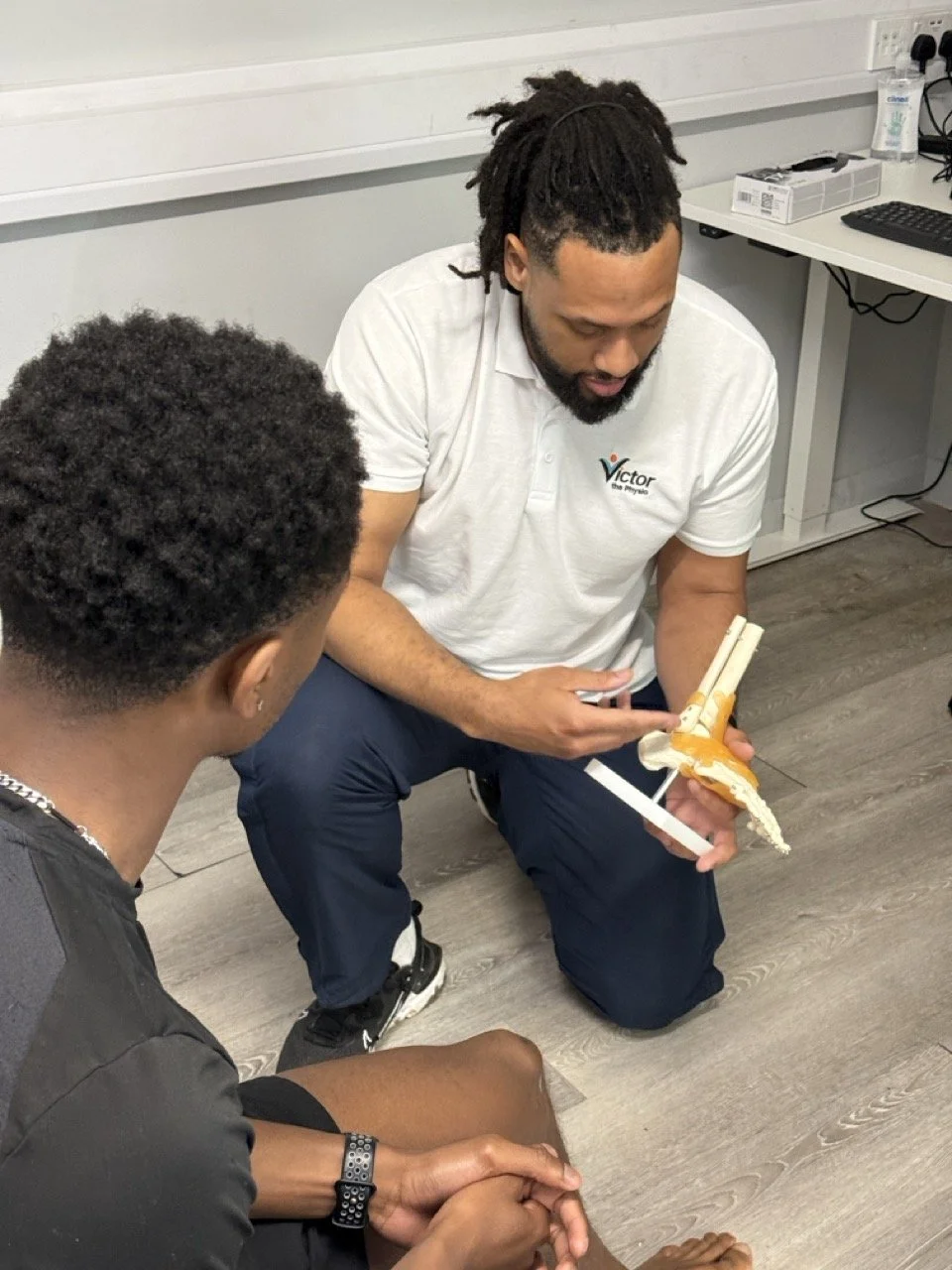 A man with dreadlocks kneeling and showing a prosthetic leg to another person in a room with a white desk and computer equipment.