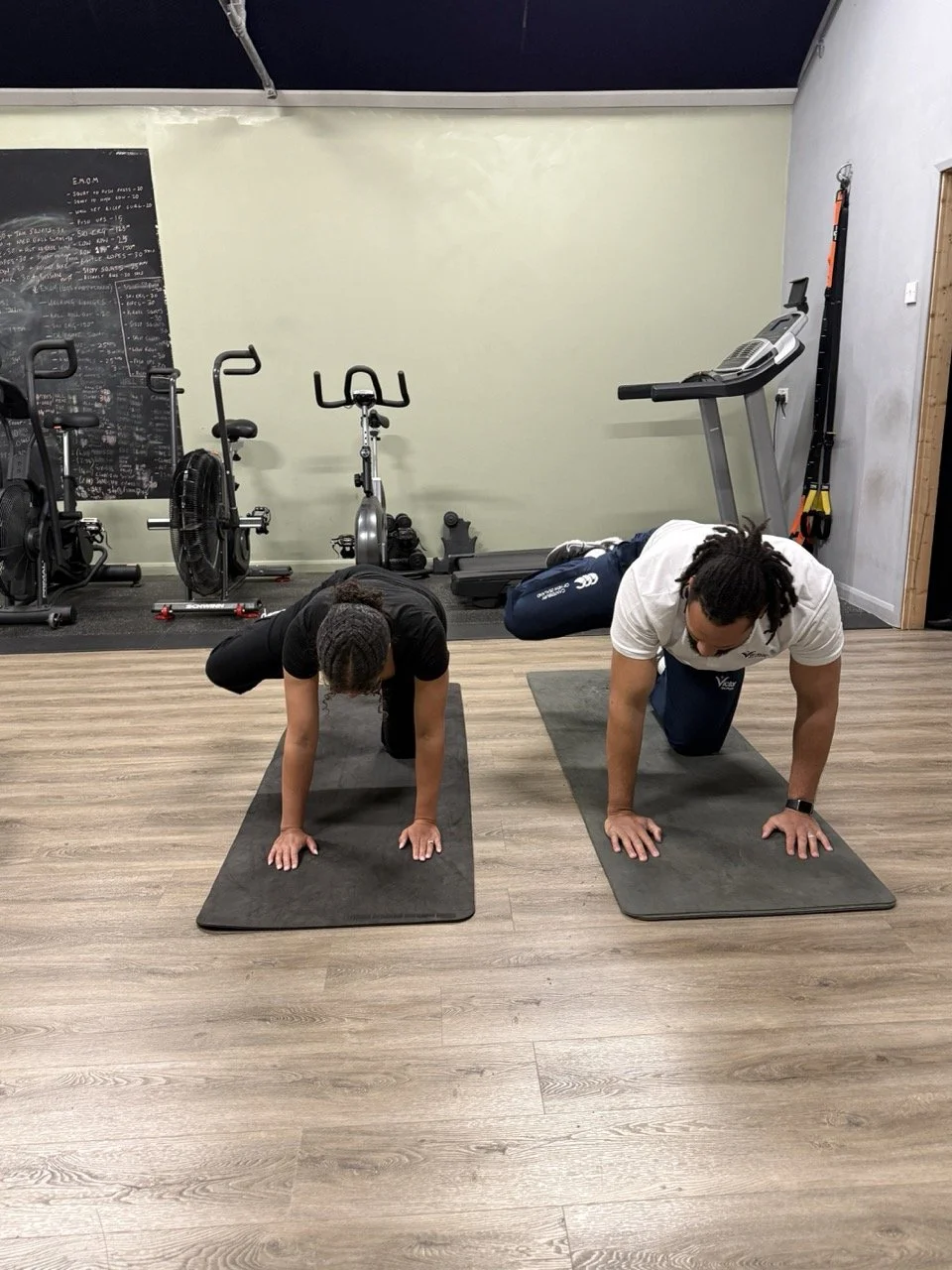 Two people practicing yoga on mats in a gym, in a child's pose position, with gym equipment in the background.
