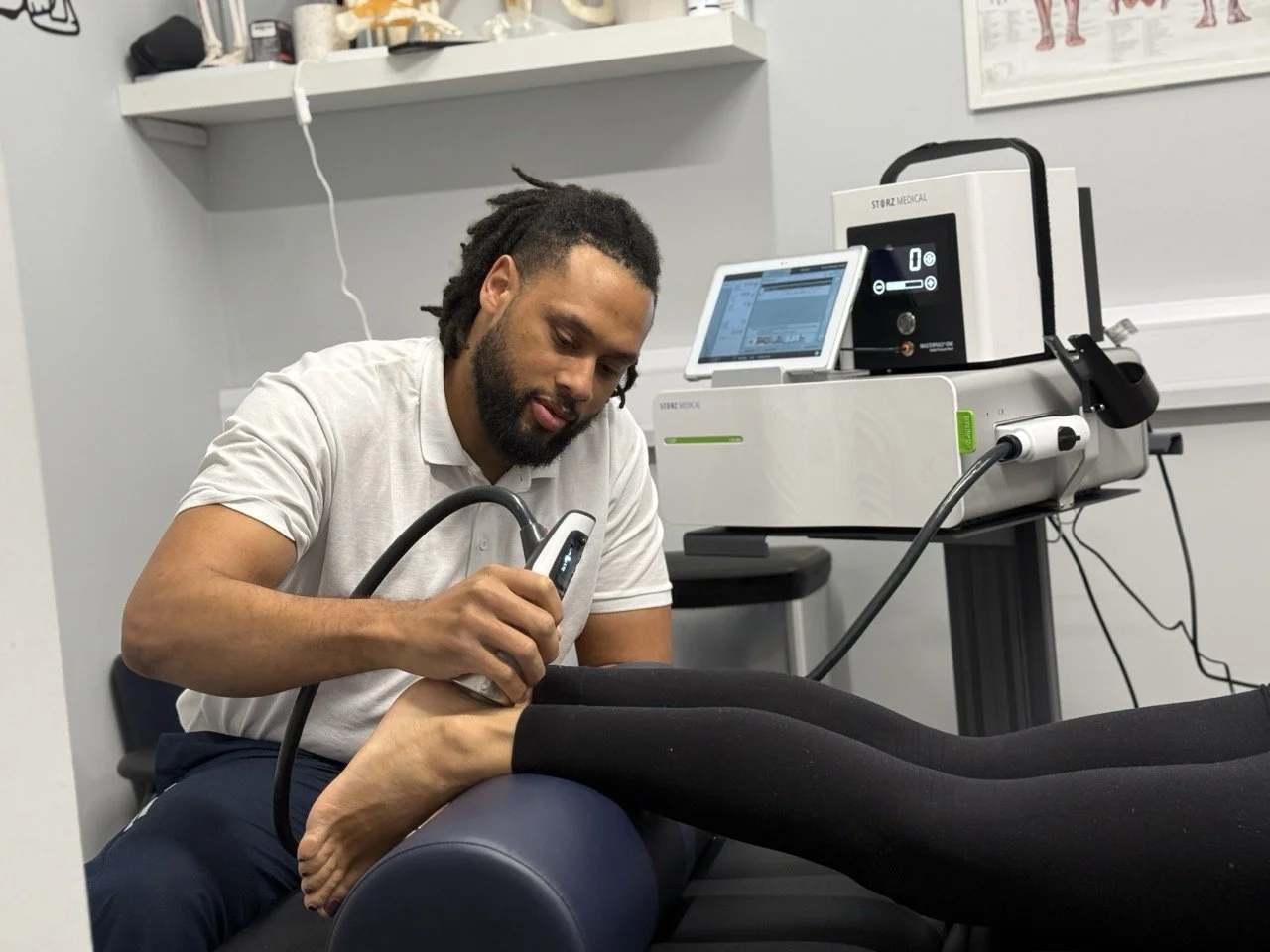 A physiotherapist performing shockwave therapy on a patient's foot using a handheld device in a clinical setting with physiotherapy equipment and a monitor in the background.