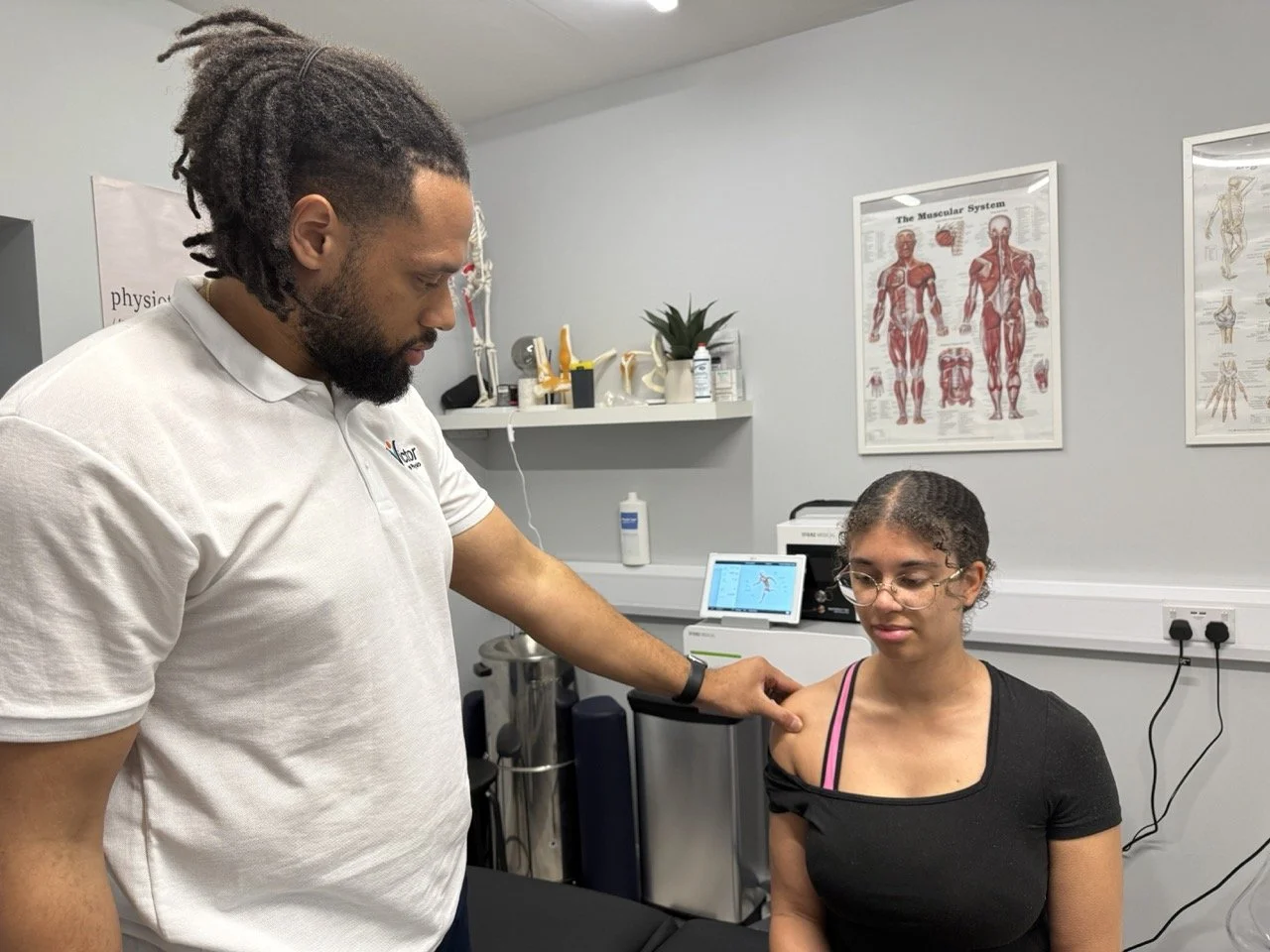 A healthcare professional examines a young woman in a medical office or clinic. The woman has curly hair, glasses, and is wearing a black shirt with pink straps. The professional is touching her shoulder and looking at her attentively. Medical posters and equipment are visible in the background.