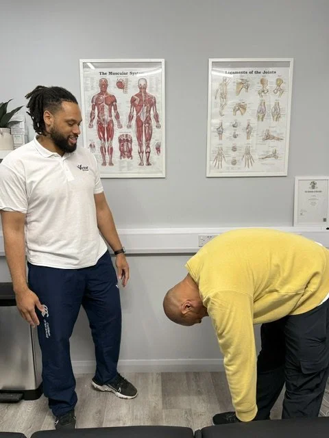 A man in a yellow sweatshirt bending over while a male physiotherapist in a white polo shirt observes.