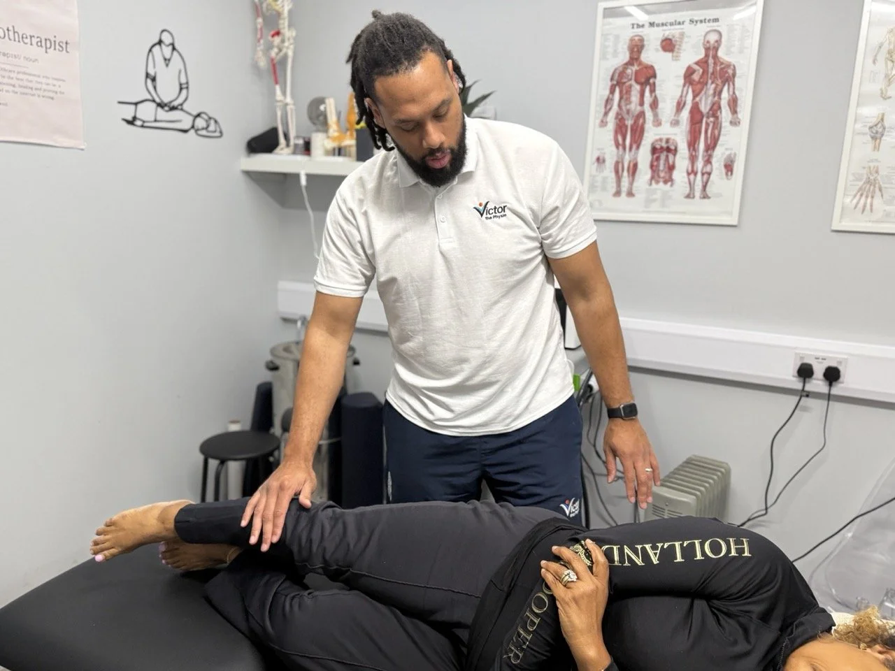 A chiropractor performing an adjustment on a woman lying face down on a treatment table in a medical office, with anatomical posters on the wall behind them.