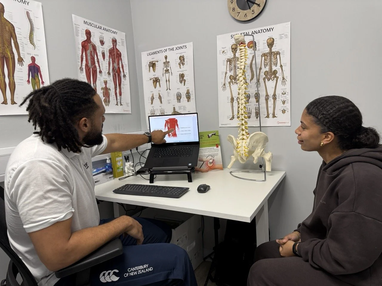 A physiotherapy consultation in an consultation with anatomy posters on the wall, a computer displaying an anatomical image of the shoulder, and a life-sized skeleton model.