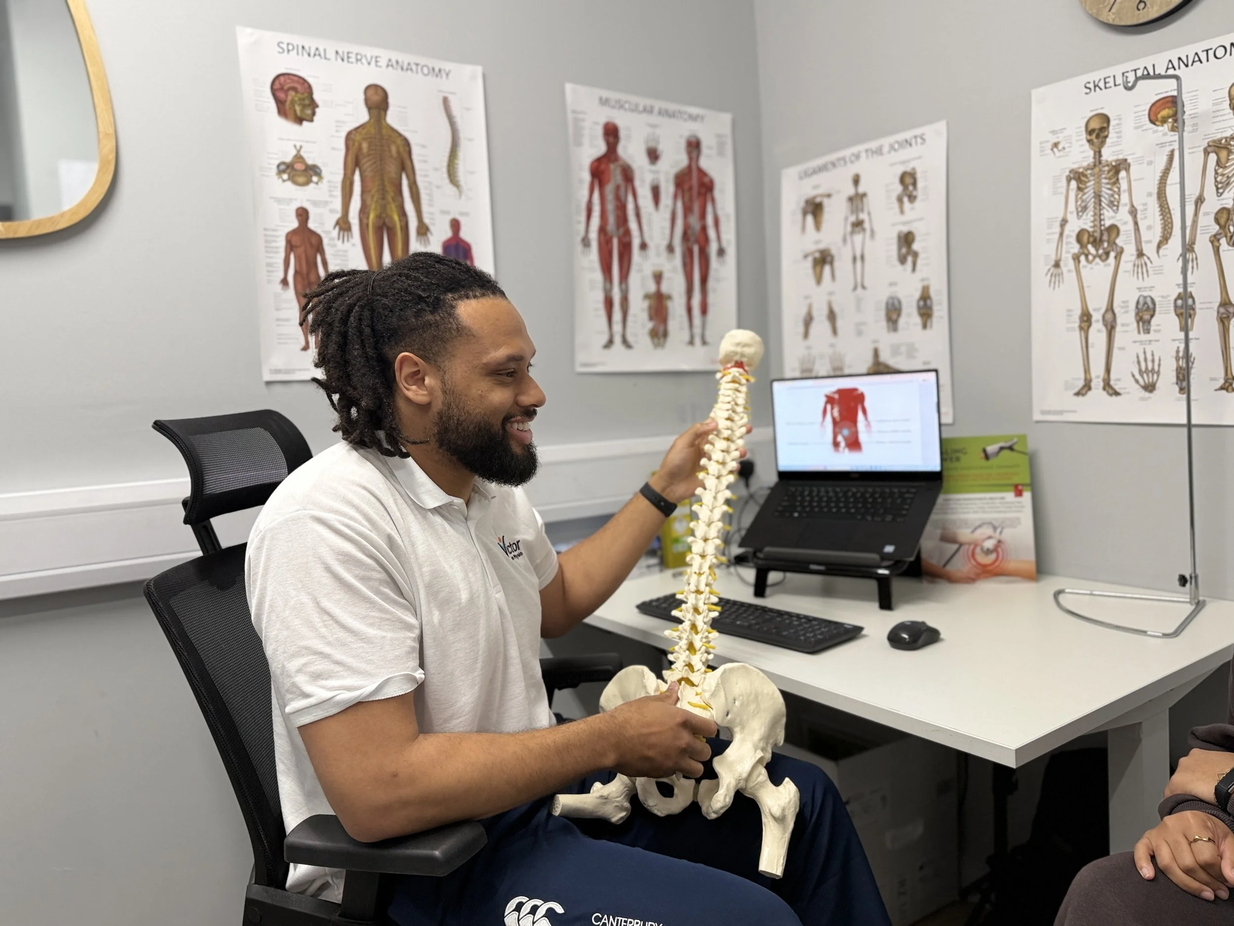 A man sitting in an office chair, smiling, is holding a model of the human spine and pelvis. Behind him, anatomical posters of the human body are pinned to the wall, including spine, muscular, skeletal, and joint diagrams. There is a computer monitor on the desk displaying a red figure of the human body.