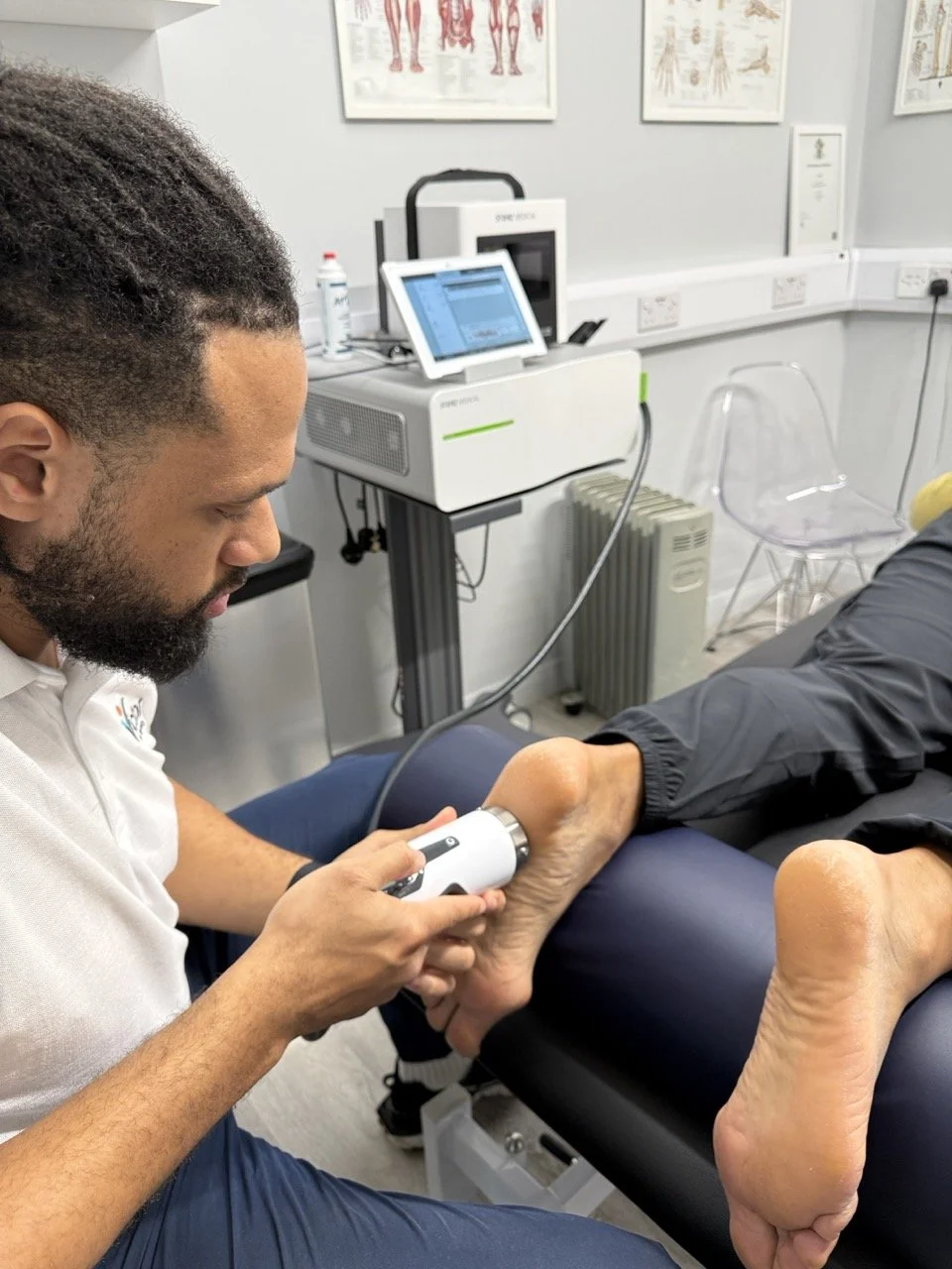A physiotherapist is using a handheld shockwave therapy device on a person's foot in a physiotherapy clinic. The room has anatomical posters, medical equipment, and a transparent chair.