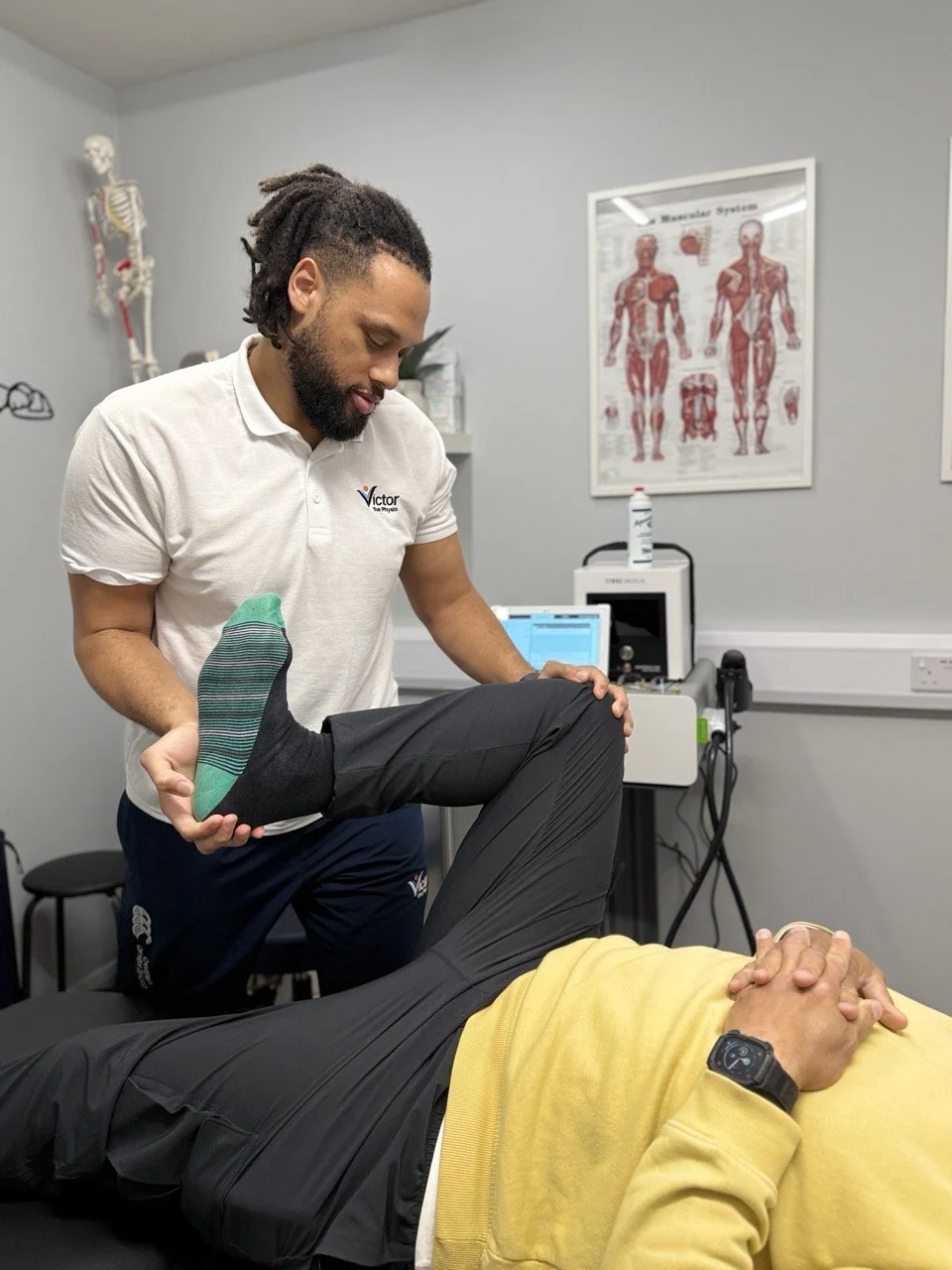 A physical therapist assesses a patient's knee joint while the patient is lying on a treatment table in a medical clinic. The therapist is holding the patient's knee, and medical posters and equipment are visible in the background.