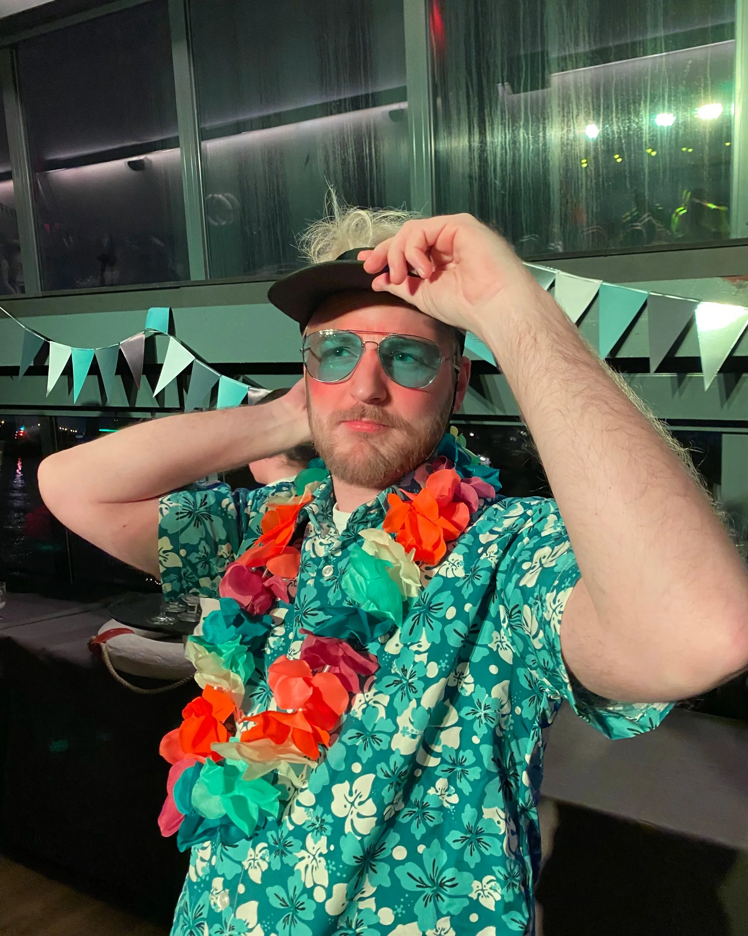 A man wearing sunglasses, a black cap, a blue Hawaiian shirt, and a colorful lei, adjusting his cap in an indoor setting decorated with hanging pennant banners and window reflections.