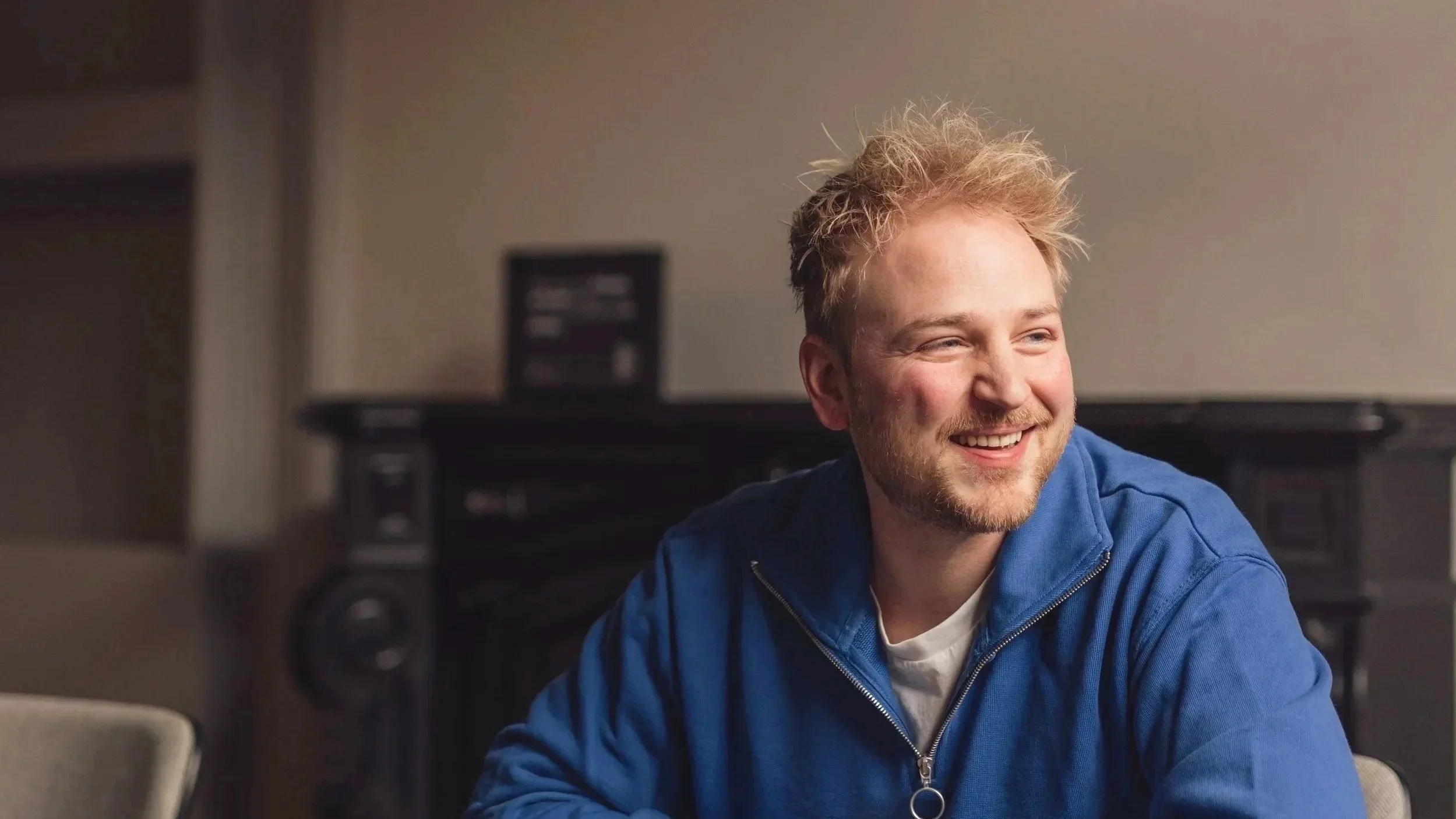 A young man with blond curly hair and a beard, smiling and wearing a blue zip-up jacket, is sitting indoors with a blurred background including speakers and furniture.