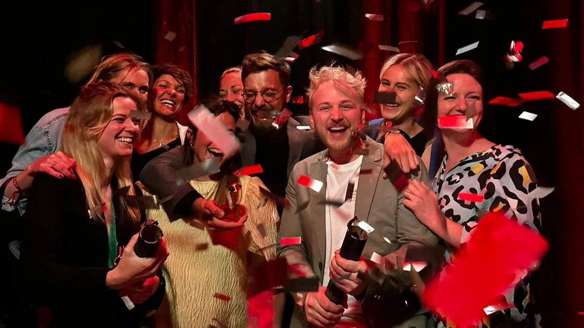 Group of nine happy people celebrating at Creative Club Belgium with confetti, some holding bottles, on a stage with red lights.