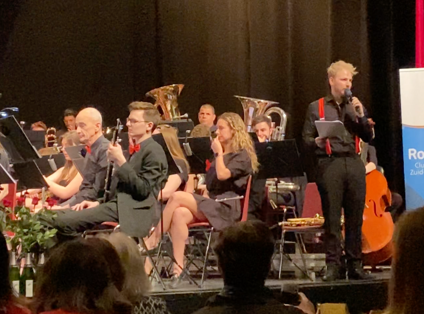 Young man in black attire with red suspenders speaking into a microphone while holding a notebook at a live orchestral concert.