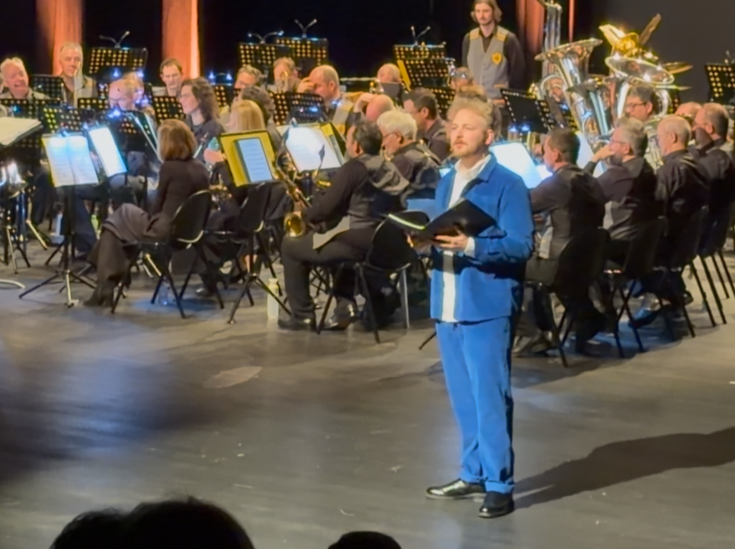 An orchestra rehearsing on stage with a presenter standing in front, holding a book, wearing a blue suit. Stage lights illuminate the scene.