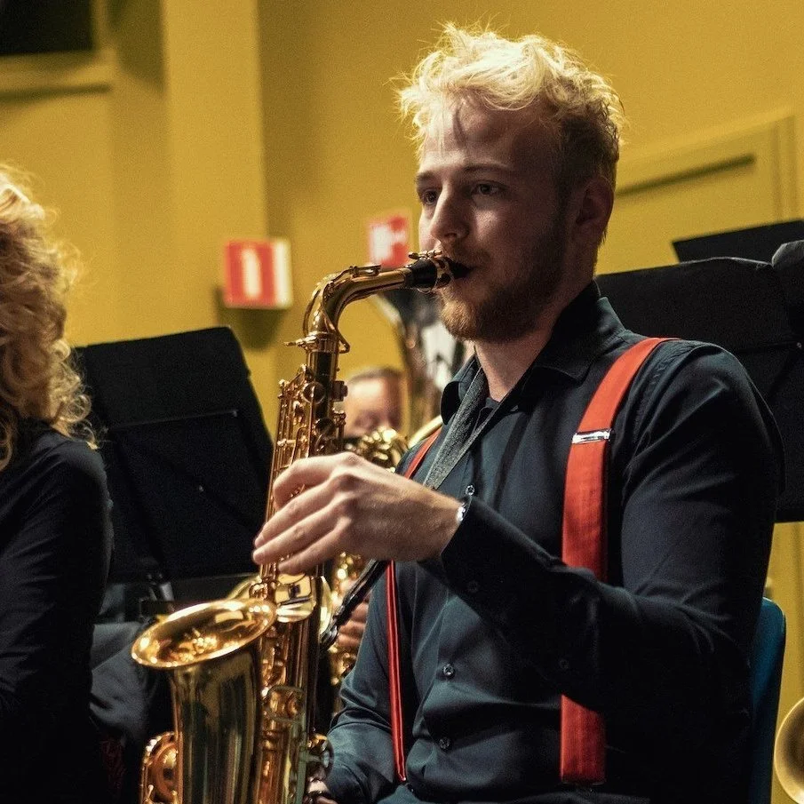 Young man with blonde, curly hair playing a saxophone in an indoor setting.