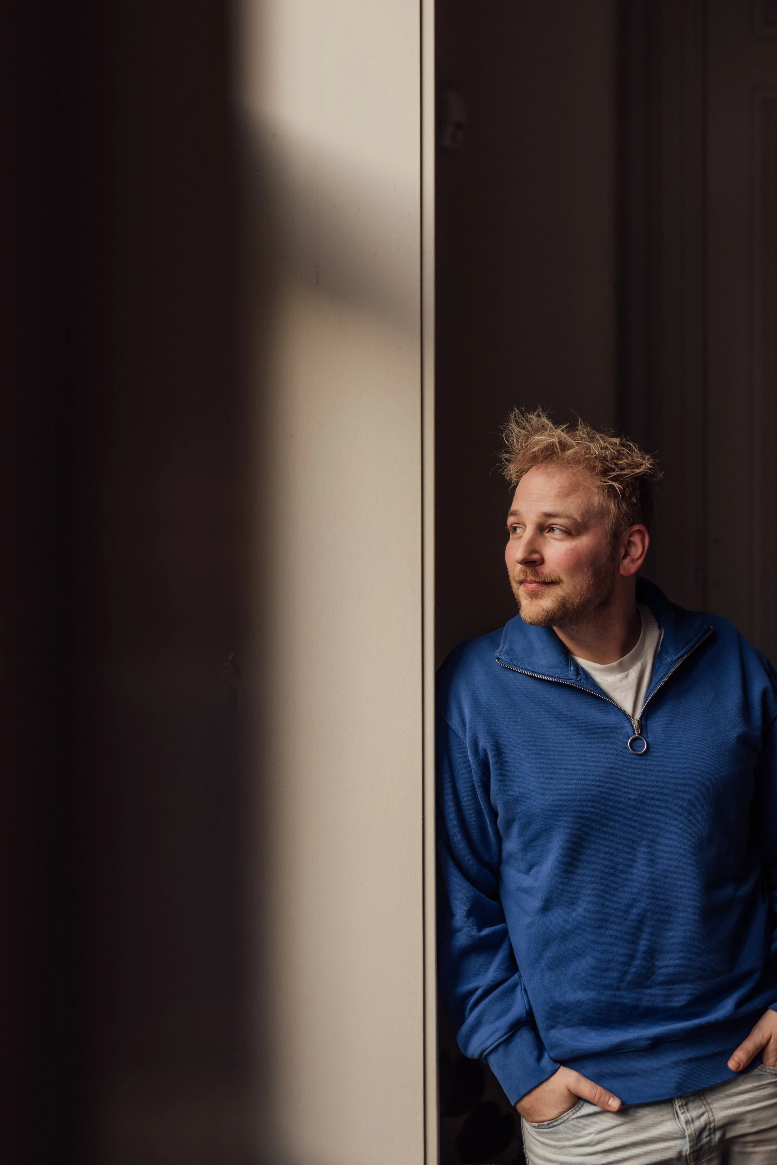 A young man with messy hair and a slight beard, wearing a blue zip-up hoodie over a white shirt, is standing against a dark wall. He is looking to his right with a relaxed expression, with one hand in his pocket.