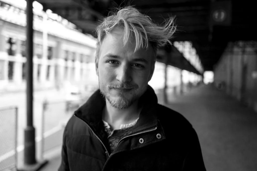 A black and white photo of a young man with messy hair and a light beard, standing at an outdoor train station platform.