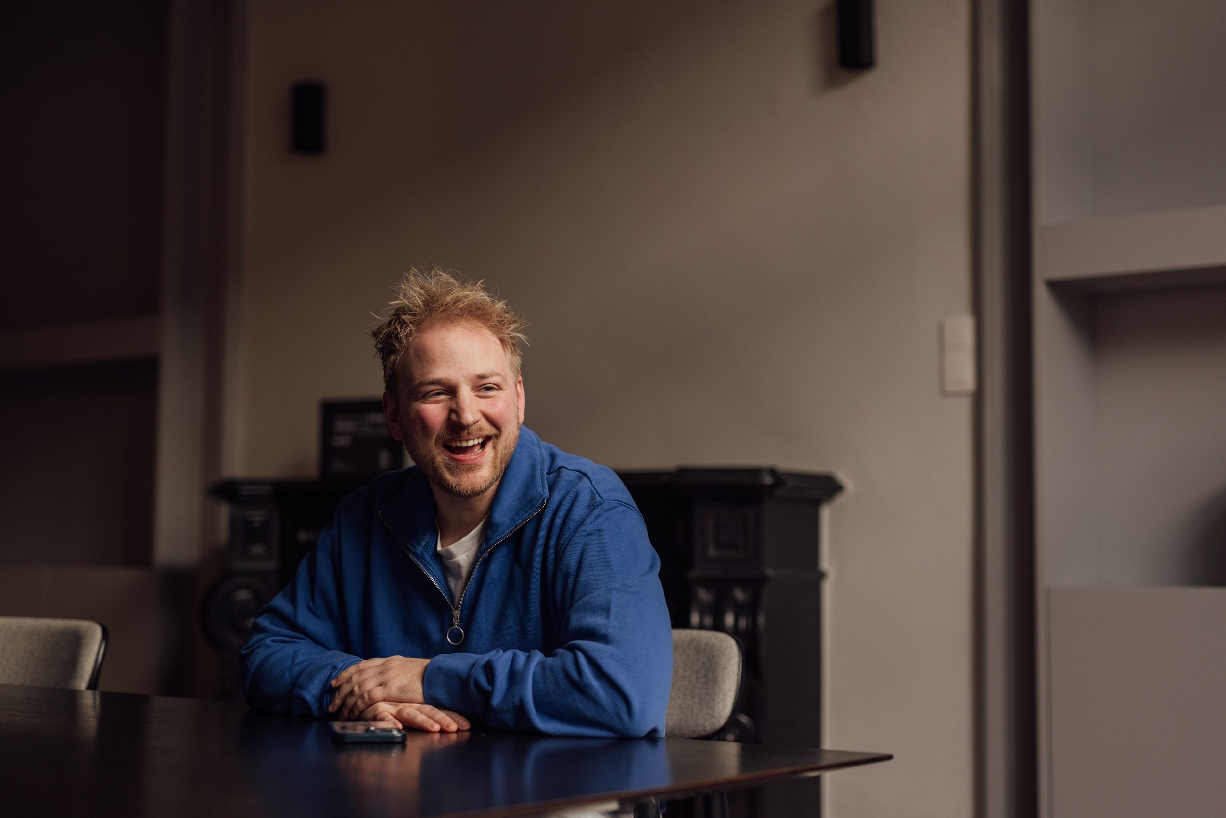 A man with blonde hair and a beard, wearing a blue jacket, sitting at a conference table, smiling and looking off to the side in a room with dark furniture and gray walls.
