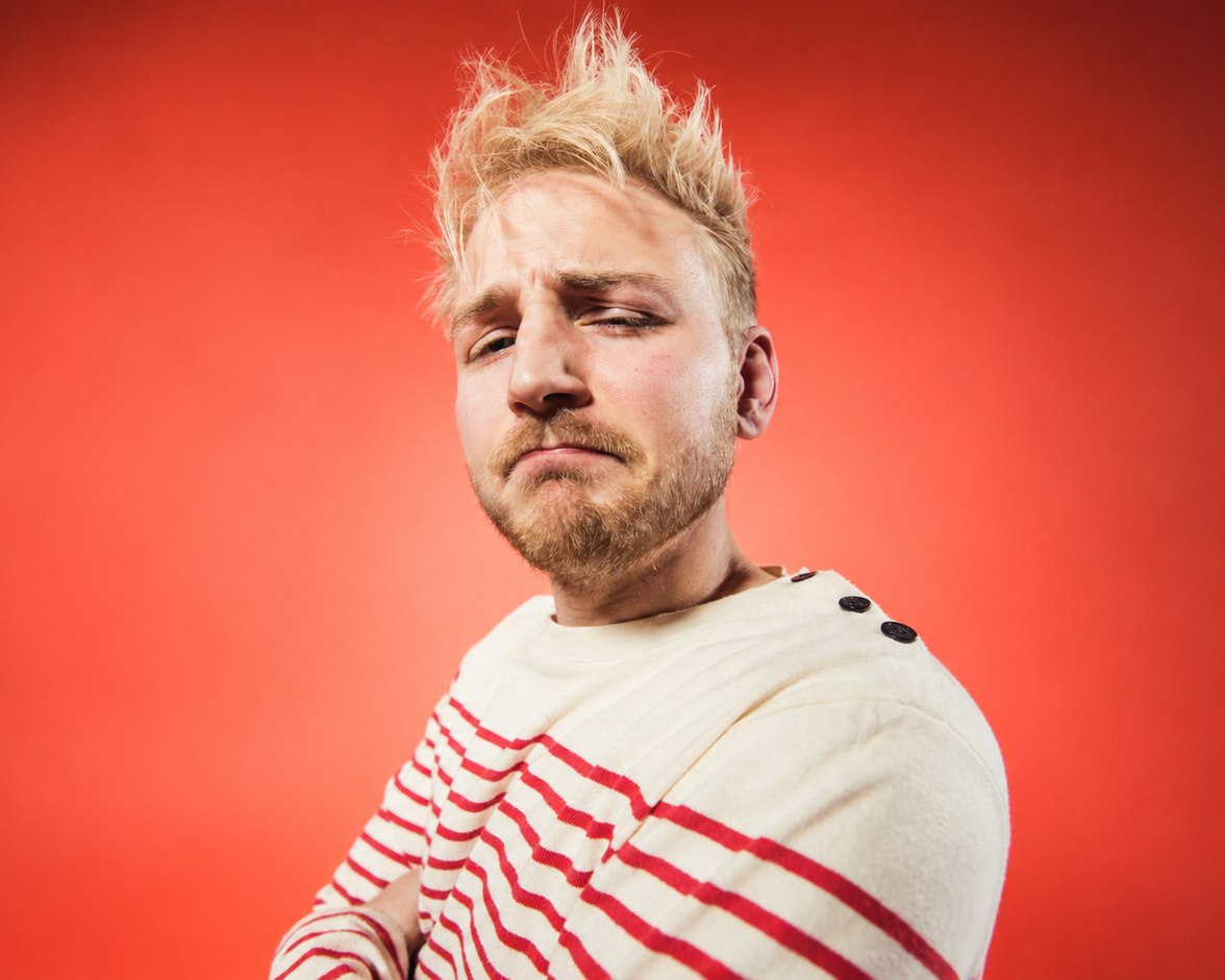 A young man with blonde, messy hair and a beard, wearing a cream-colored sweater with red and white stripes and black buttons on the shoulder, stands against a solid red background, looking at the camera with a slightly skeptical look.