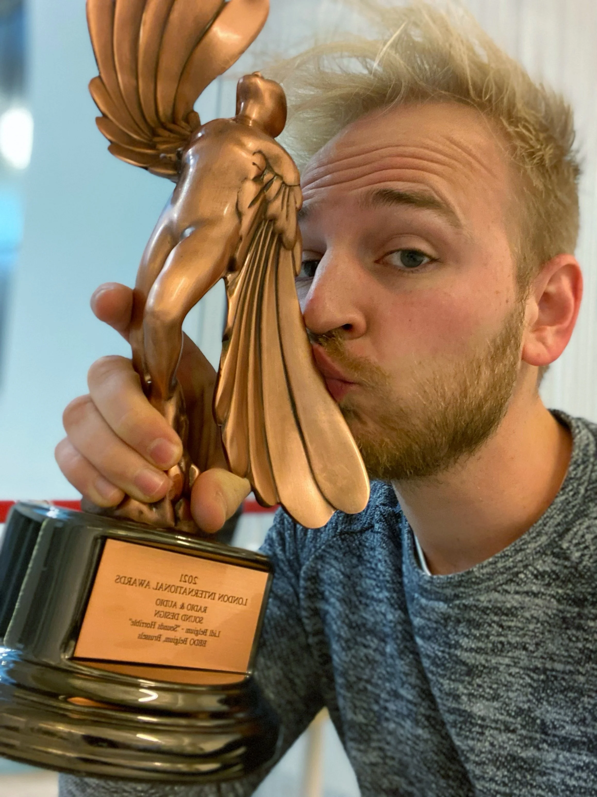 A man with blonde hair and a beard holding a London International Award , with a certificate in the foreground. The focus is on the trophy and the person's face.