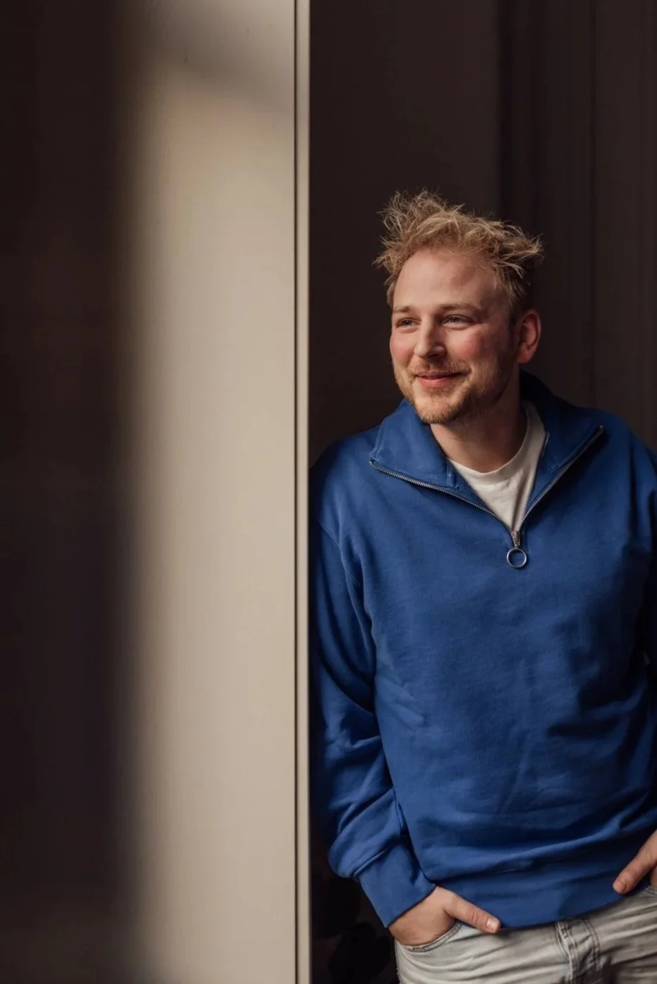 A young man with messy hair and a beard smiling and looking to his right, wearing a blue hoodie, standing next to a reflective surface.