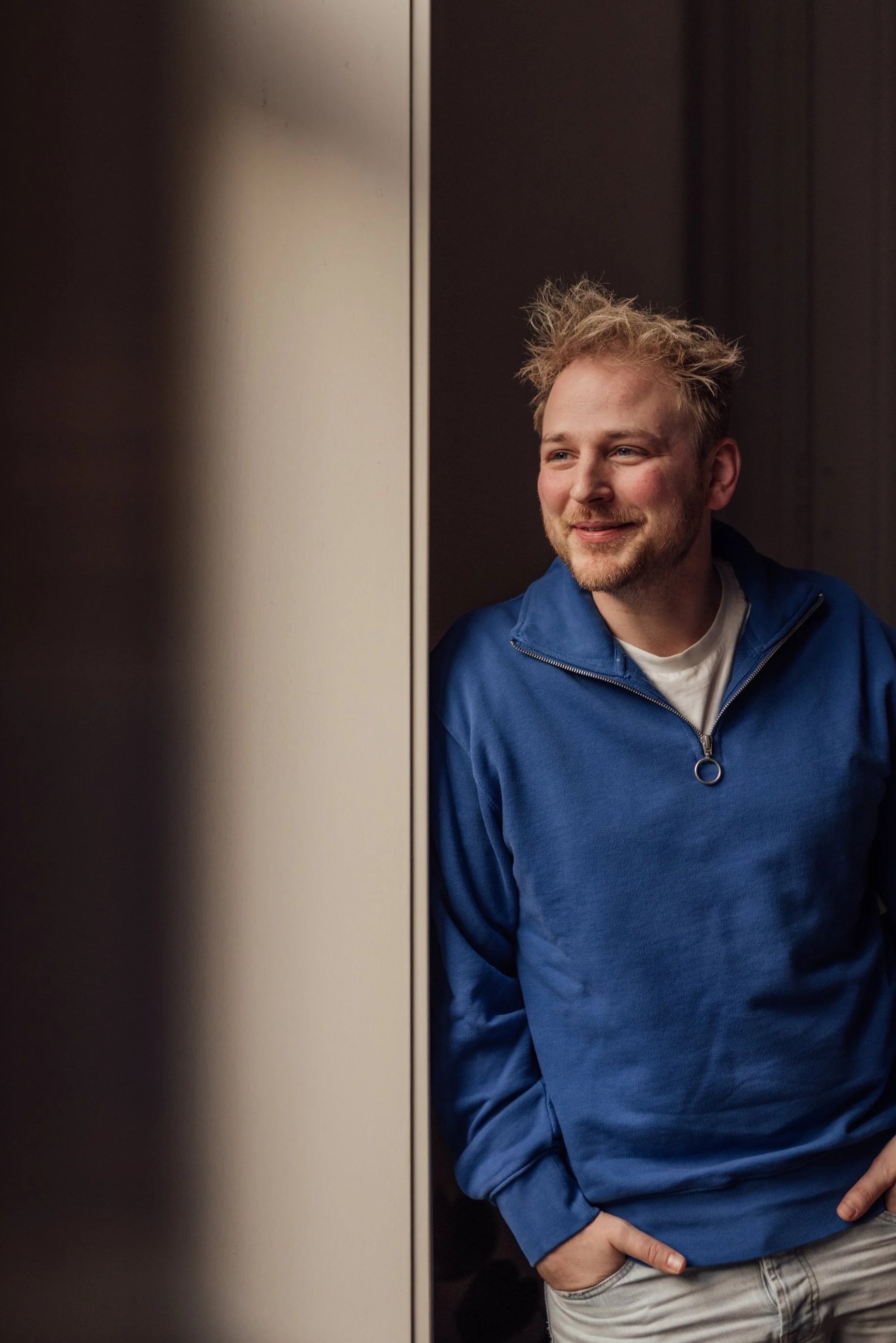 Photo of a man with messy blonde hair, wearing a blue jacket, smiling and looking to his right, with one hand in his pocket, standing in front of an elevator door.