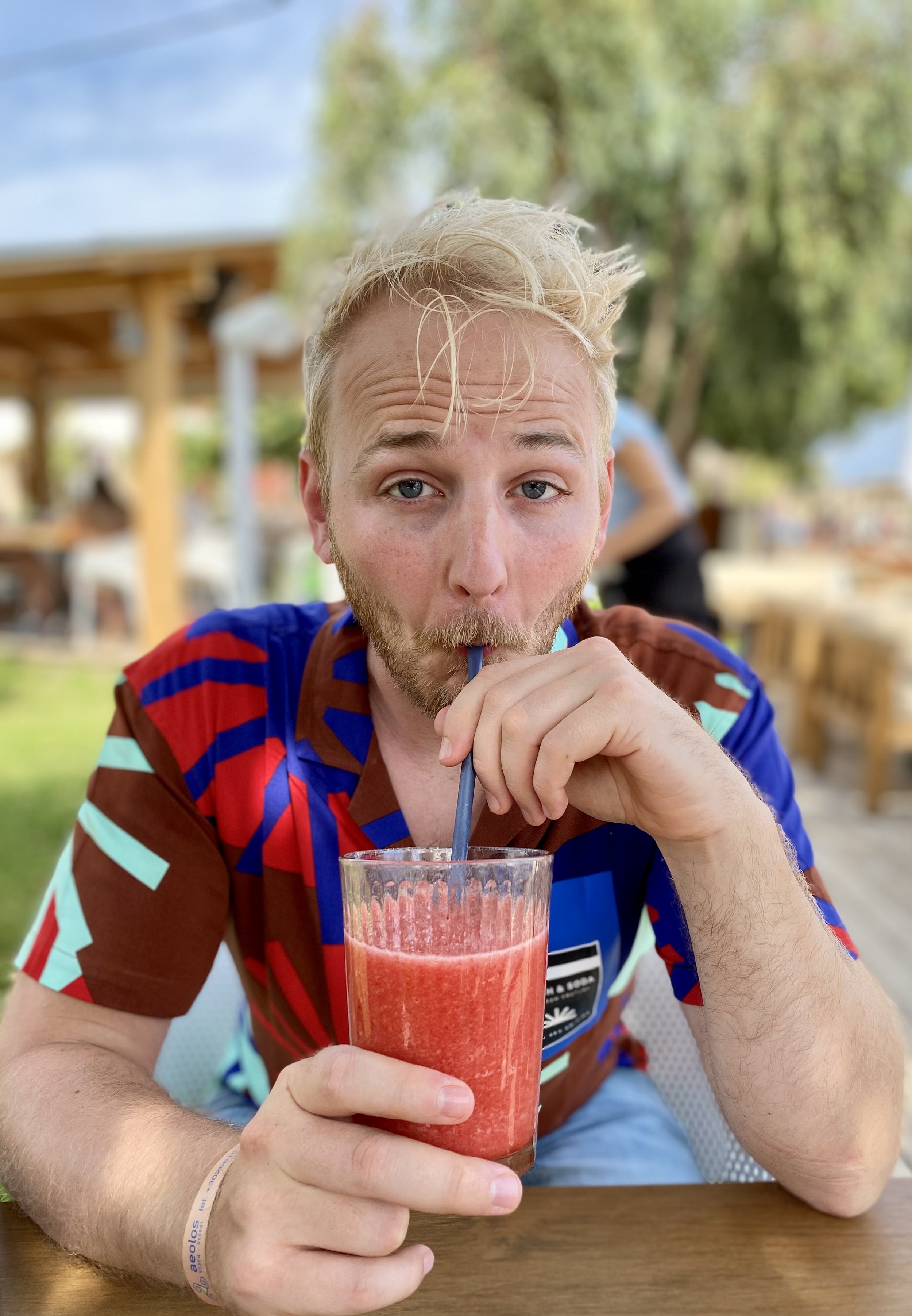 A man with blonde hair and a beard is sitting outdoors at a wooden table, drinking a pinkish-red smoothie through a straw, while looking at the camera. He is wearing a colorful striped shirt, and there are blurred green trees and a wooden structure i